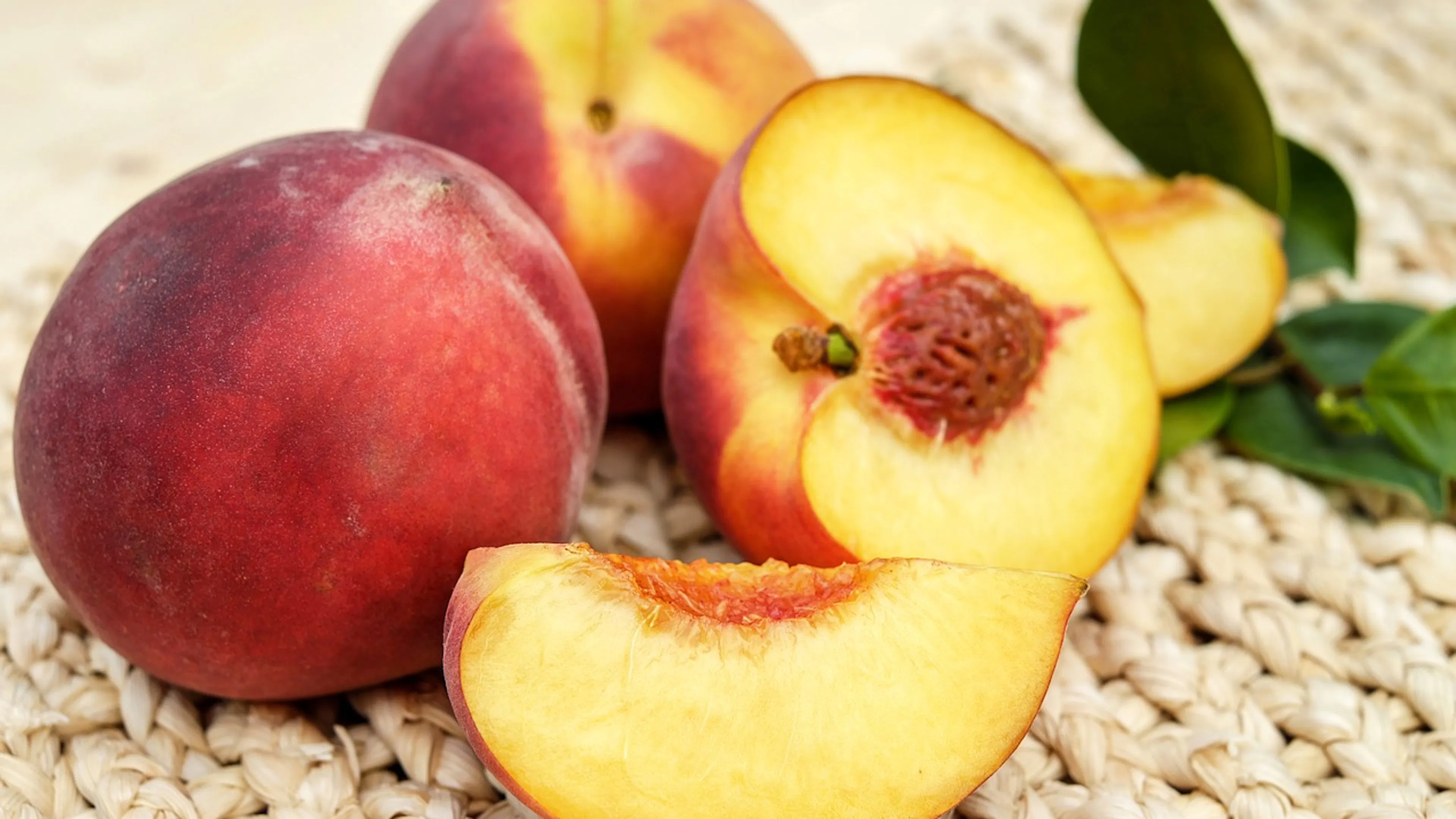 Two whole peaches resting on a woven mat and another peach cut in half in front of them