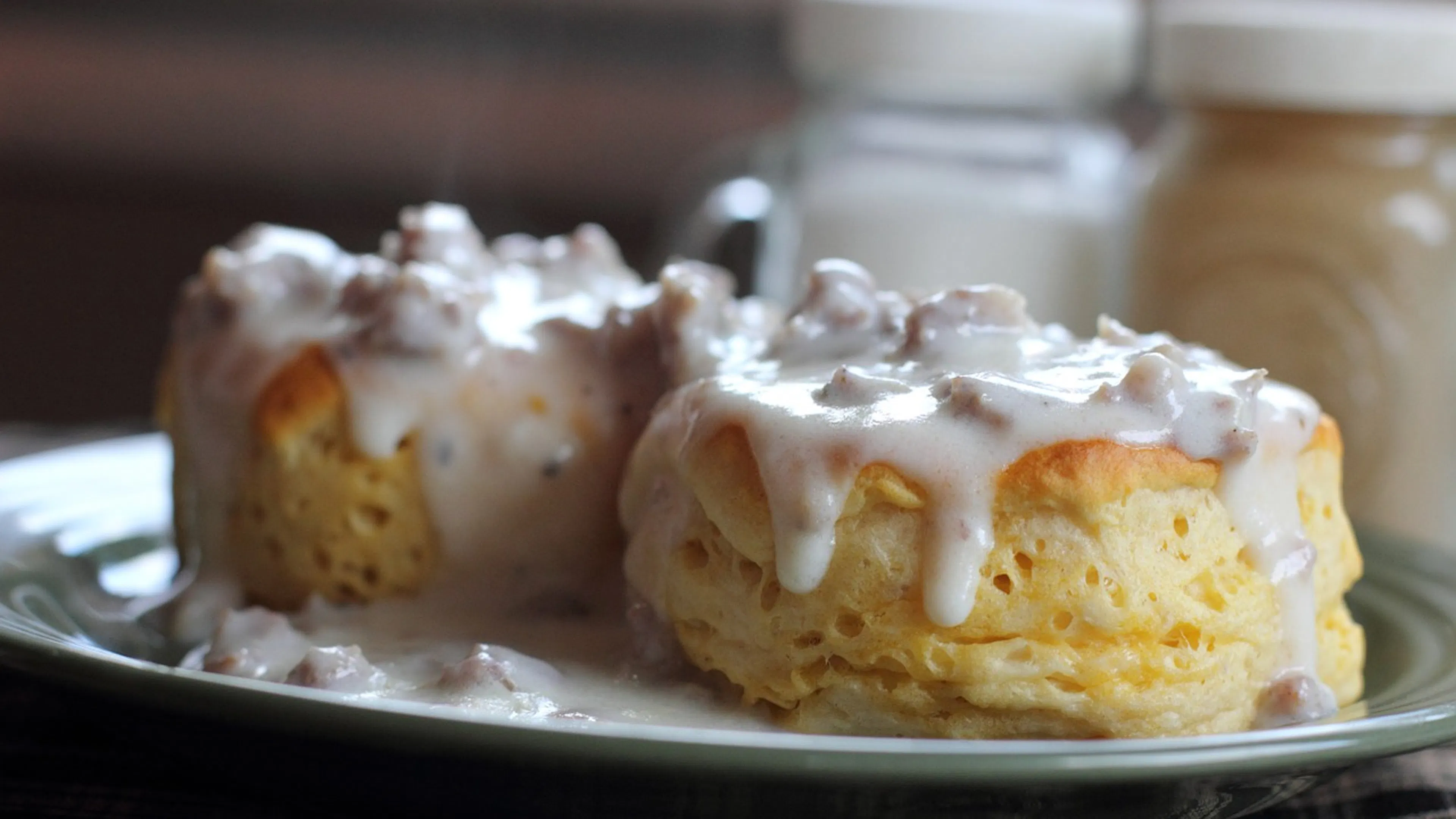 Two golden buttermilk biscuits on a small blue plate with steaming country style gravy poured over it