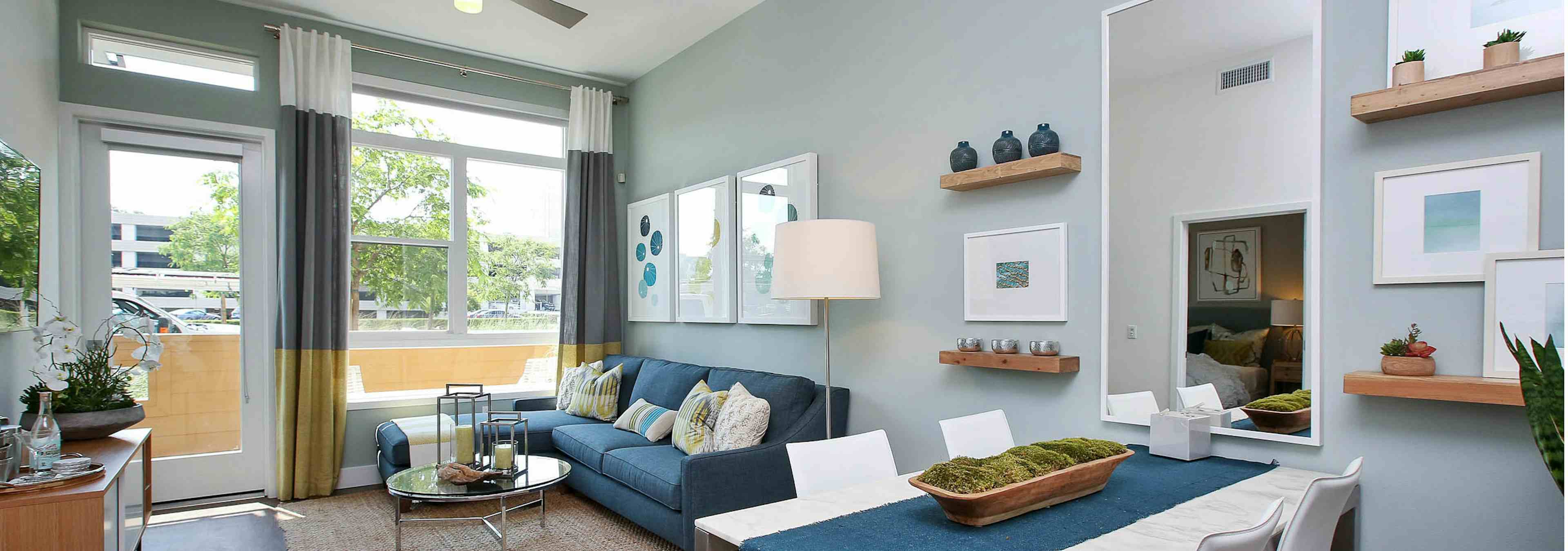 Interior view of AMLI Uptown Orange furnished apartment living room and dining area with blue couch in front of large windows