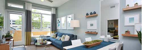 Interior view of AMLI Uptown Orange furnished apartment living room and dining area with blue couch in front of large windows