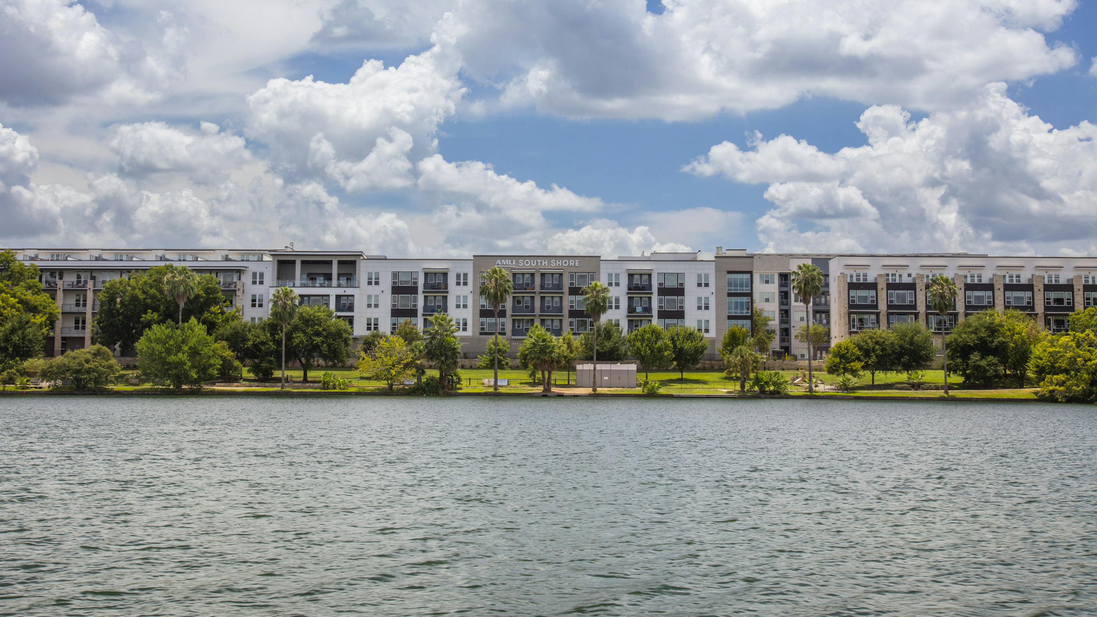 Daytime view of AMLI South Shore apartment building exterior with cloudy blue sky and lush greenery from across the water