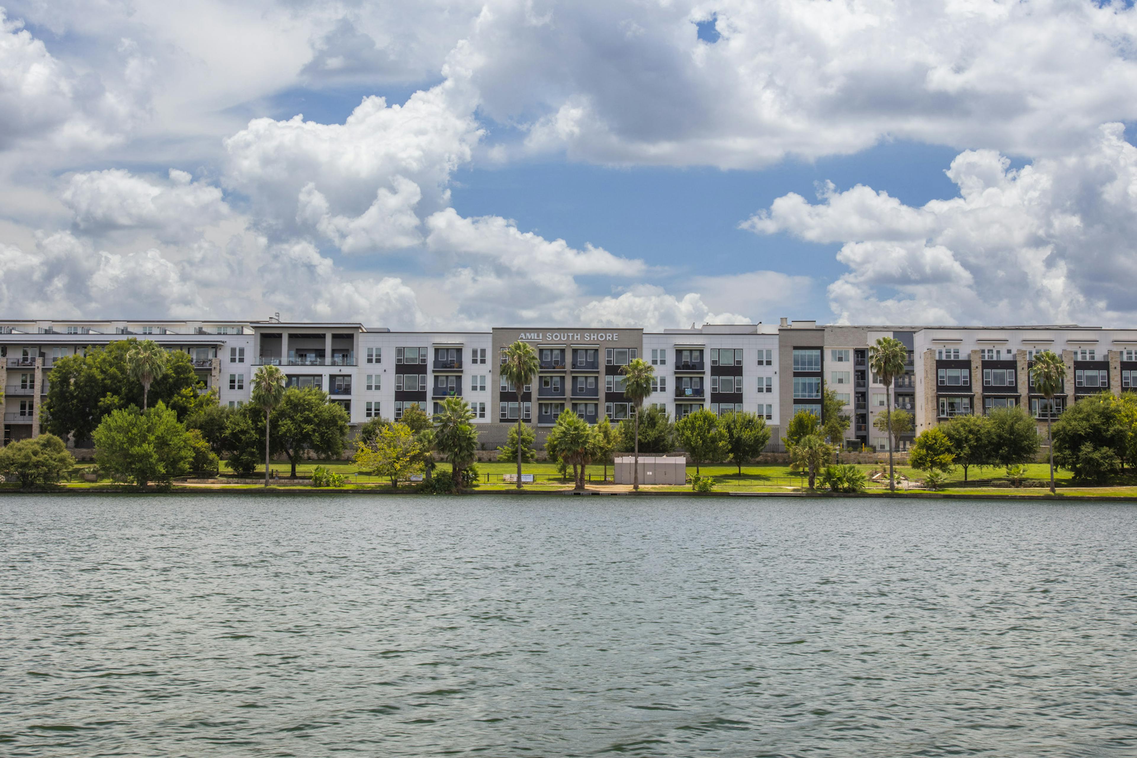 Daytime view of AMLI South Shore apartment building exterior with cloudy blue sky and lush greenery from across the water