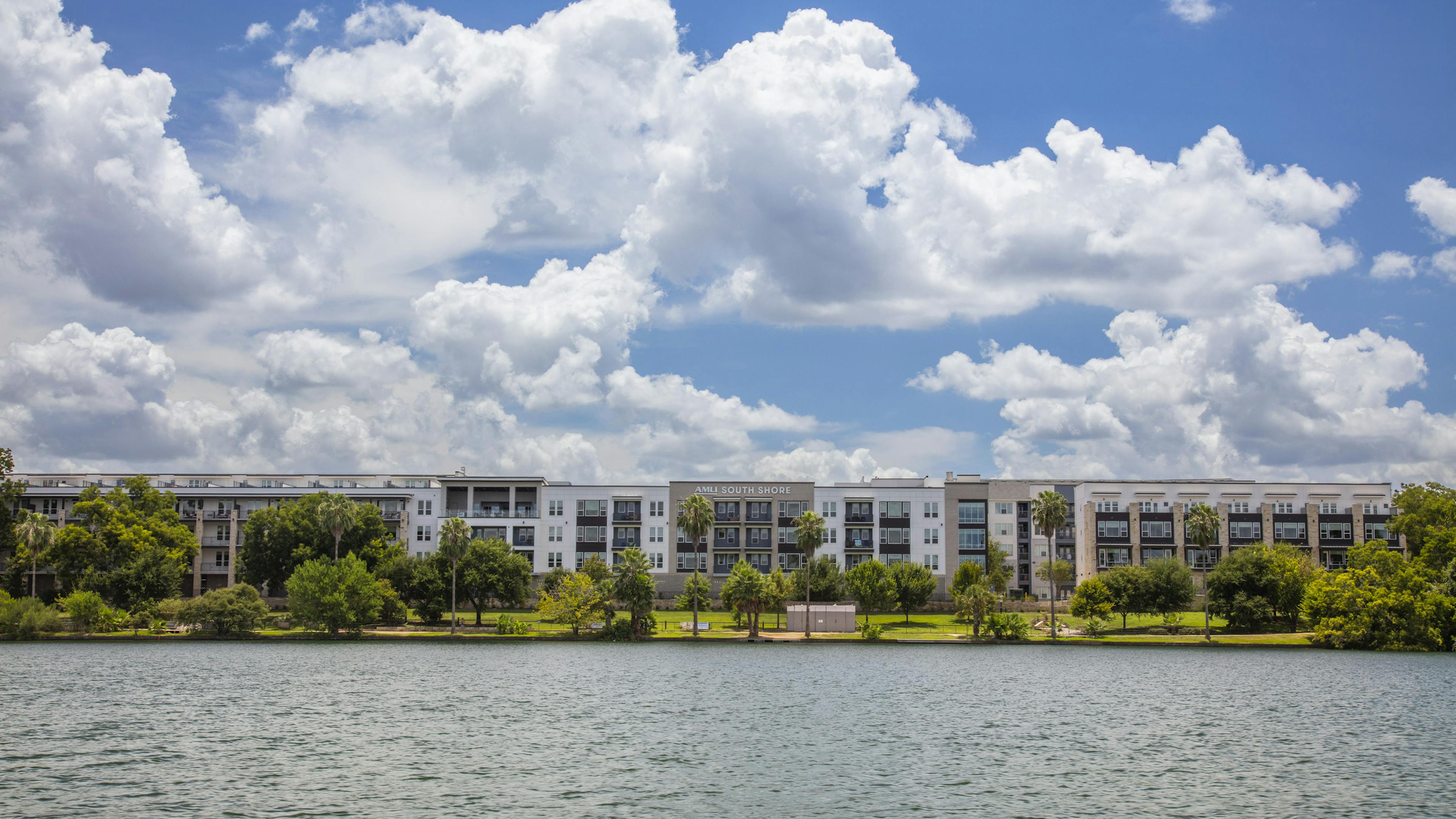 Daytime view of AMLI South Shore apartment building exterior with cloudy blue sky and lush greenery from across the water
