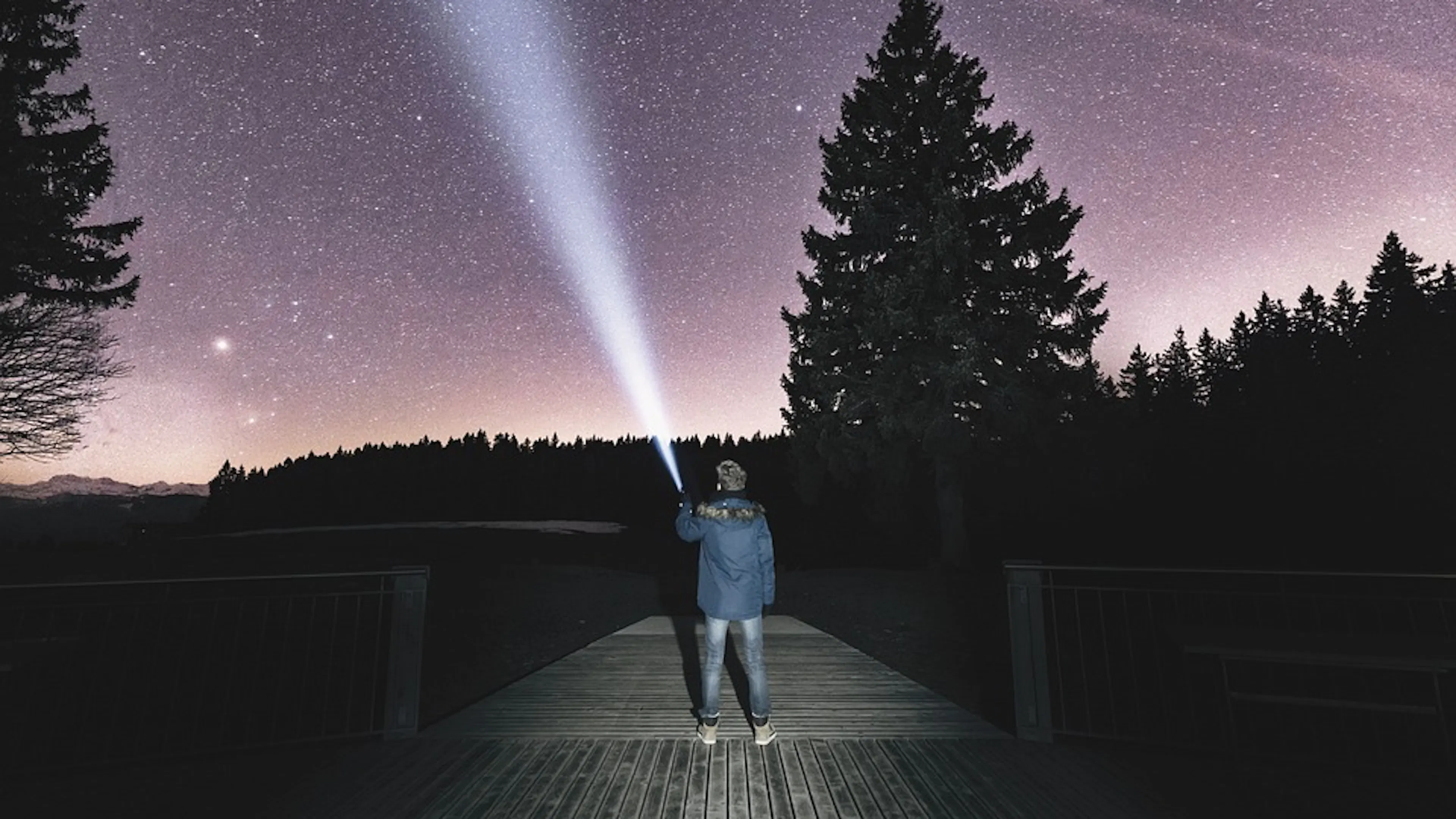 A person holding a flashlight looking up at the starry night sky standing on a pier