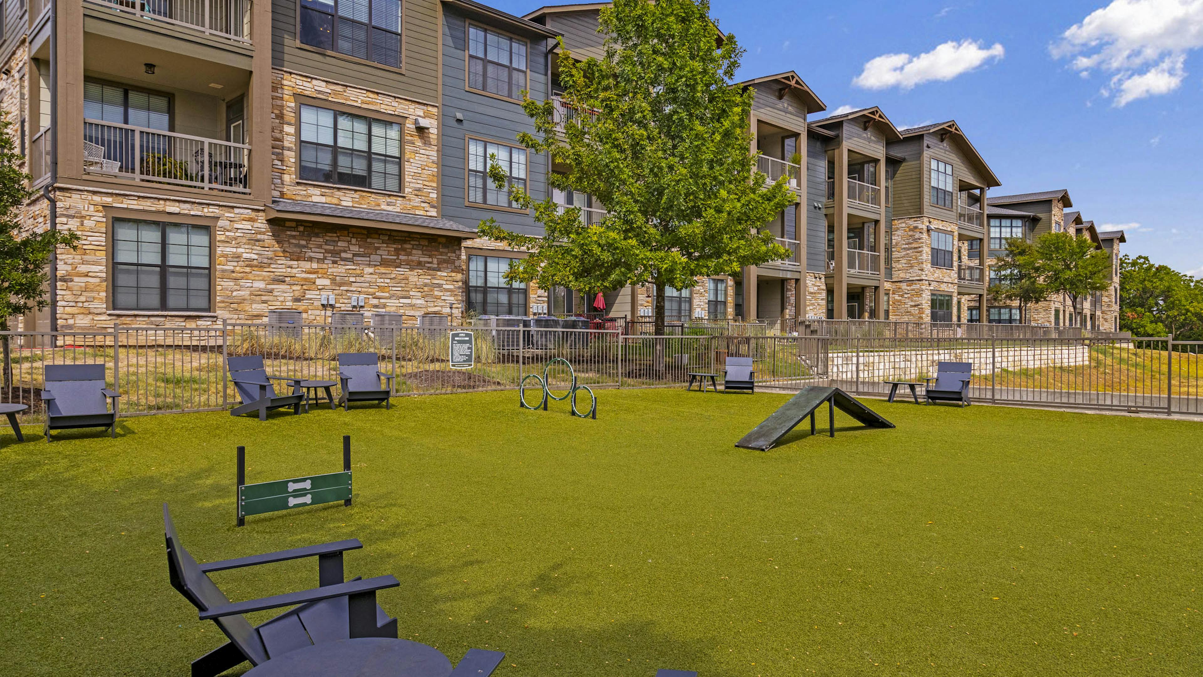 AMLI Covered Bridge dog park with green grass and black Adirondack chairs with tables and stacked stone buildings 
