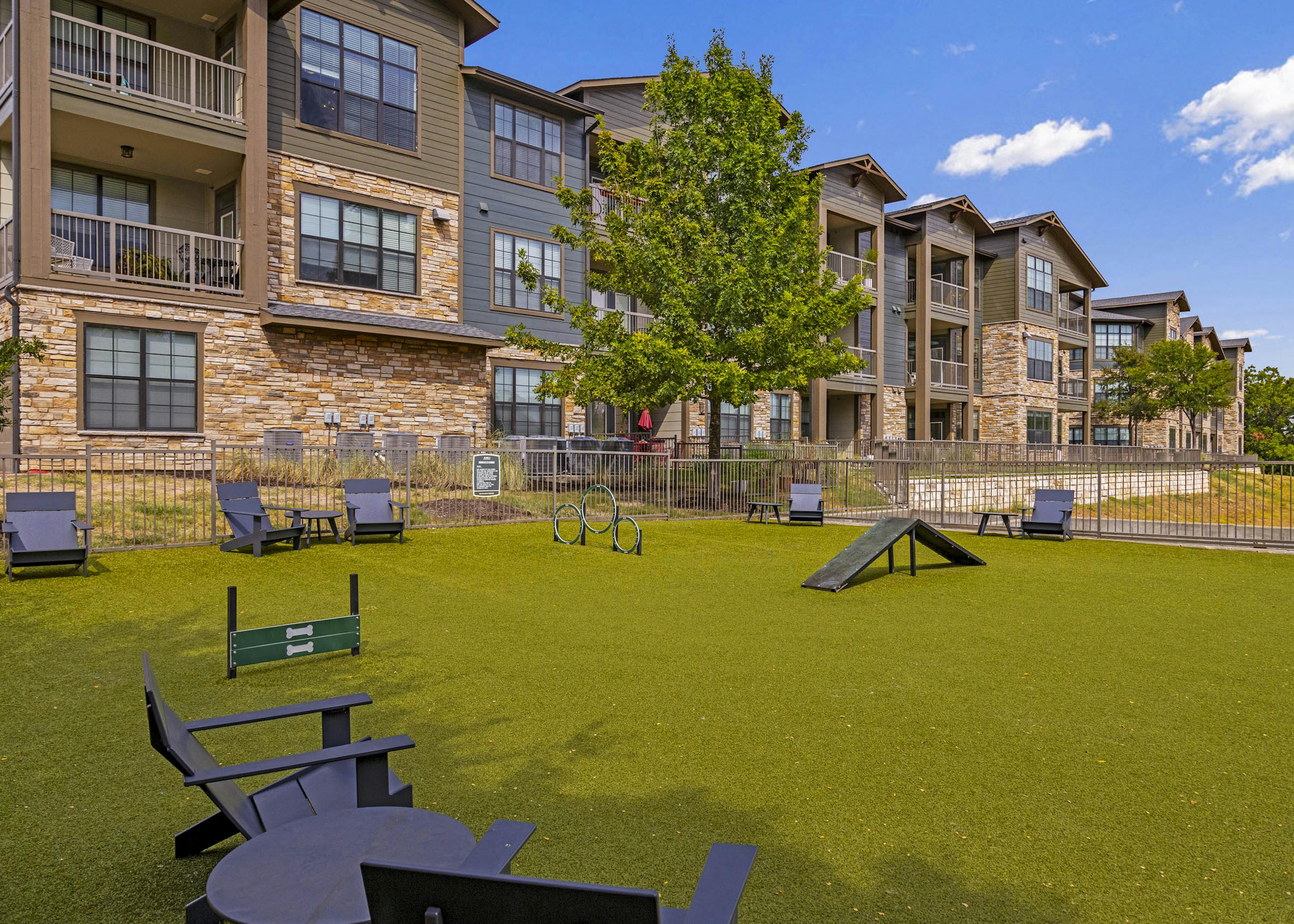 AMLI Covered Bridge dog park with green grass and black Adirondack chairs with tables and stacked stone buildings 