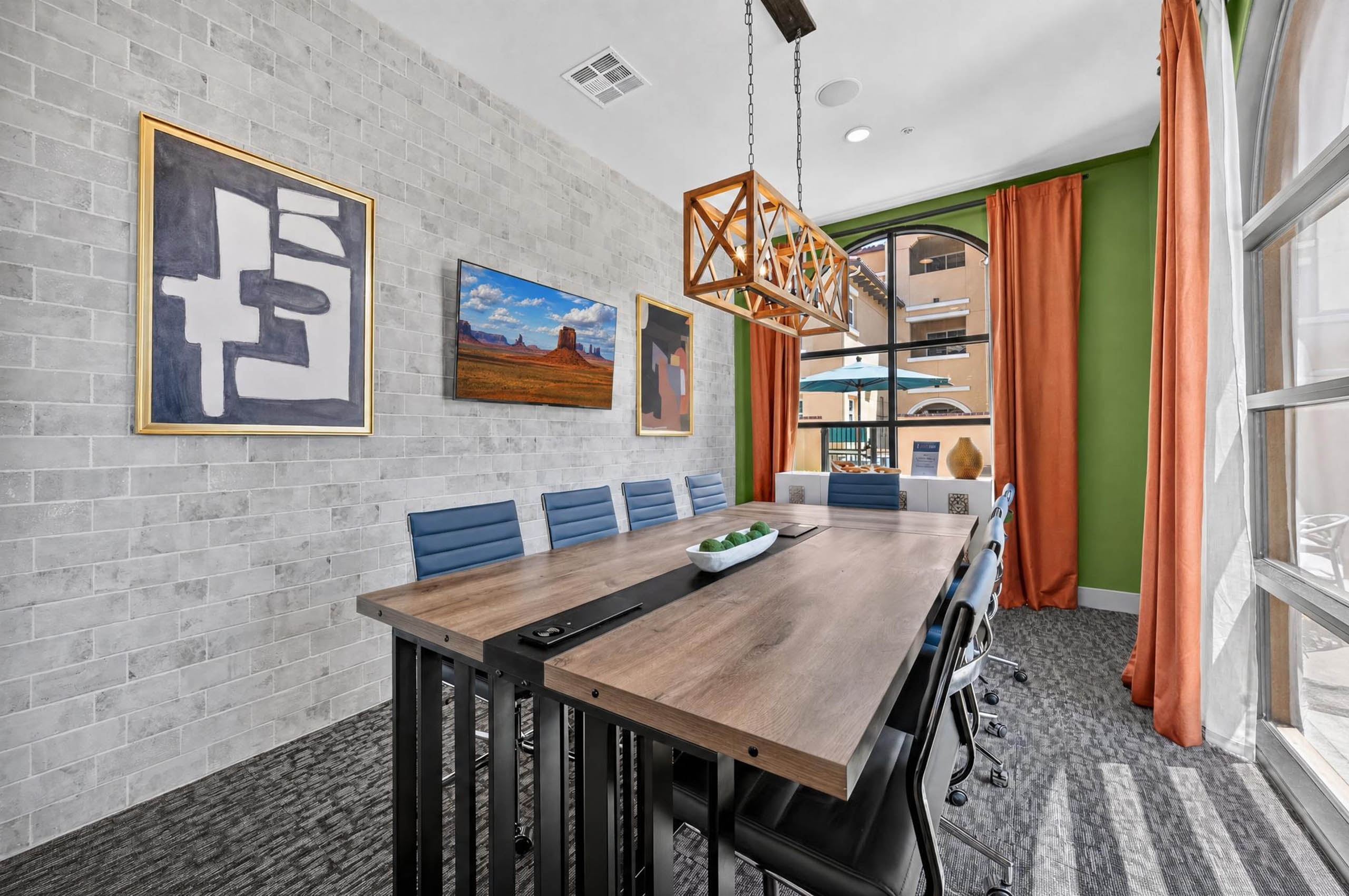 Conference room at AMLI Camarillo apartments with long wood squared table, blue chairs, framed artwork and orange curtains.