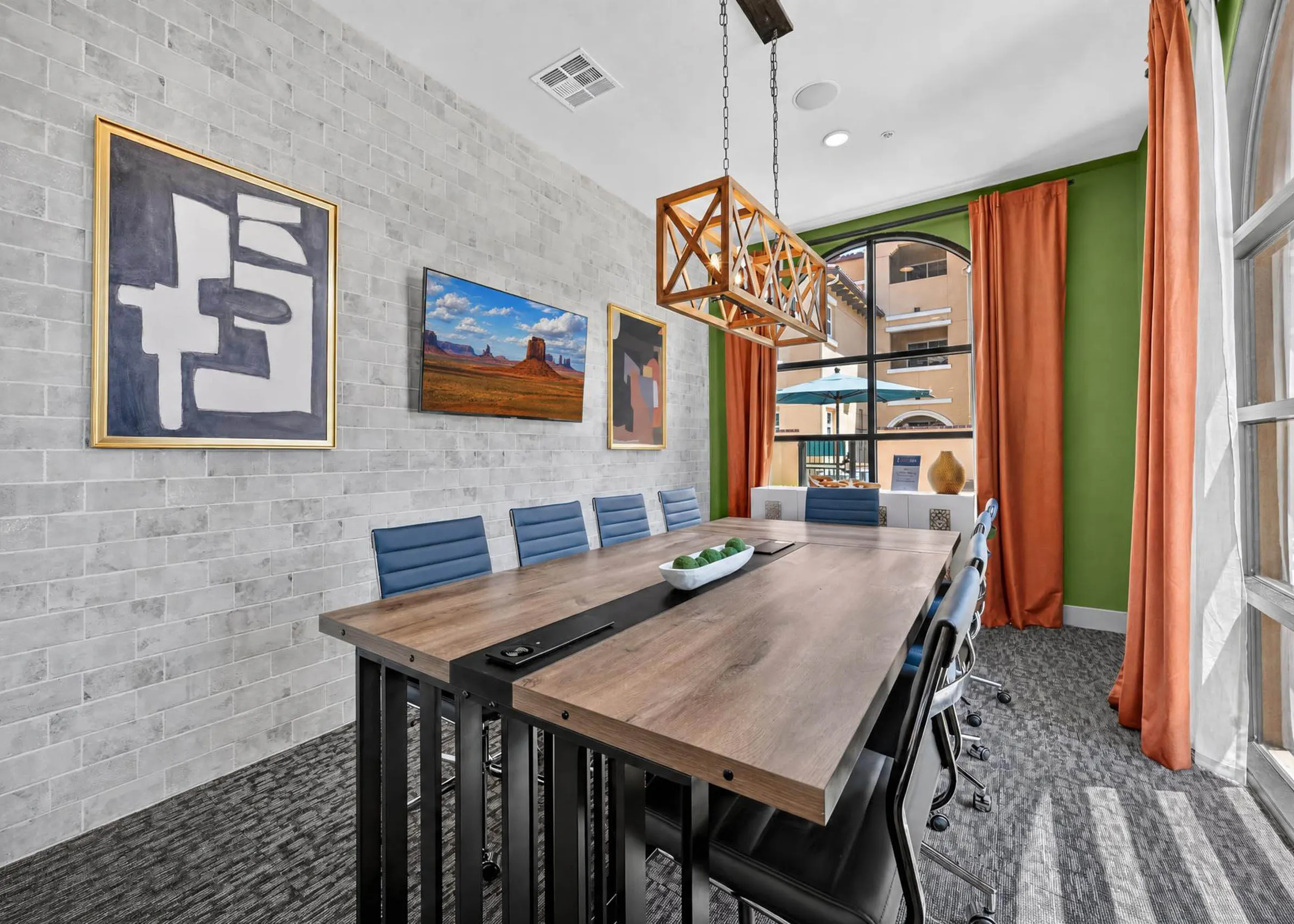 Conference room at AMLI Camarillo apartments with long wood squared table, blue chairs, framed artwork and orange curtains.