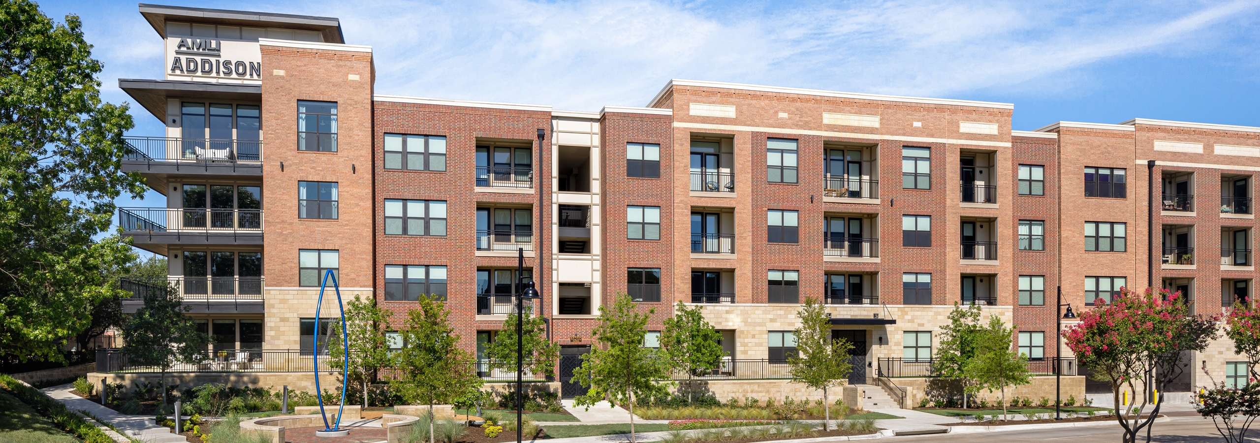 AMLI Addison apartment building with red brick and black balconies surrounded by green trees and blue sculpture in front