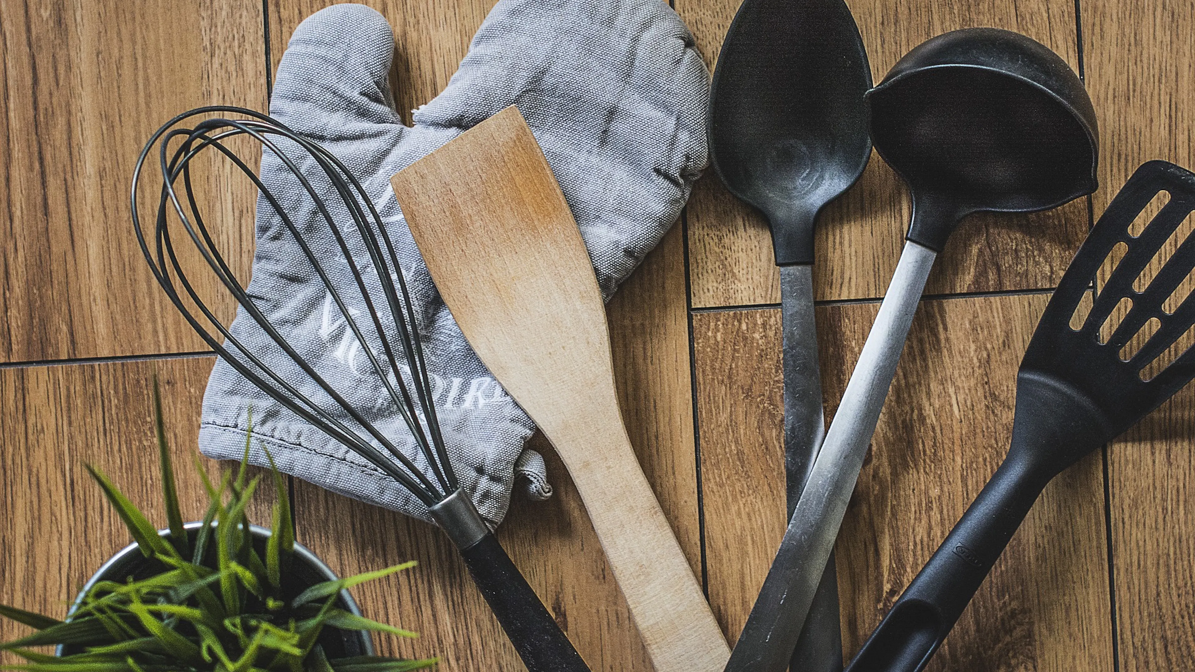 A grey oven glove on a wooden table with a whisk and a wooden spatula and plastic spoons and a spatula