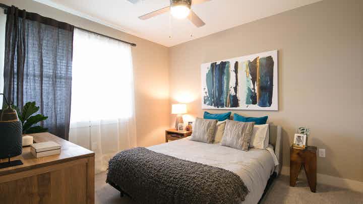 A bedroom at AMLI Dry Creek apartments with gray and blue linens and a dressers with plants and a painting and large window