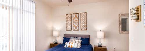 A bedroom at AMLI Arista apartments showcasing a bed with blue sheets and artwork hanging and a window with blinds drawn