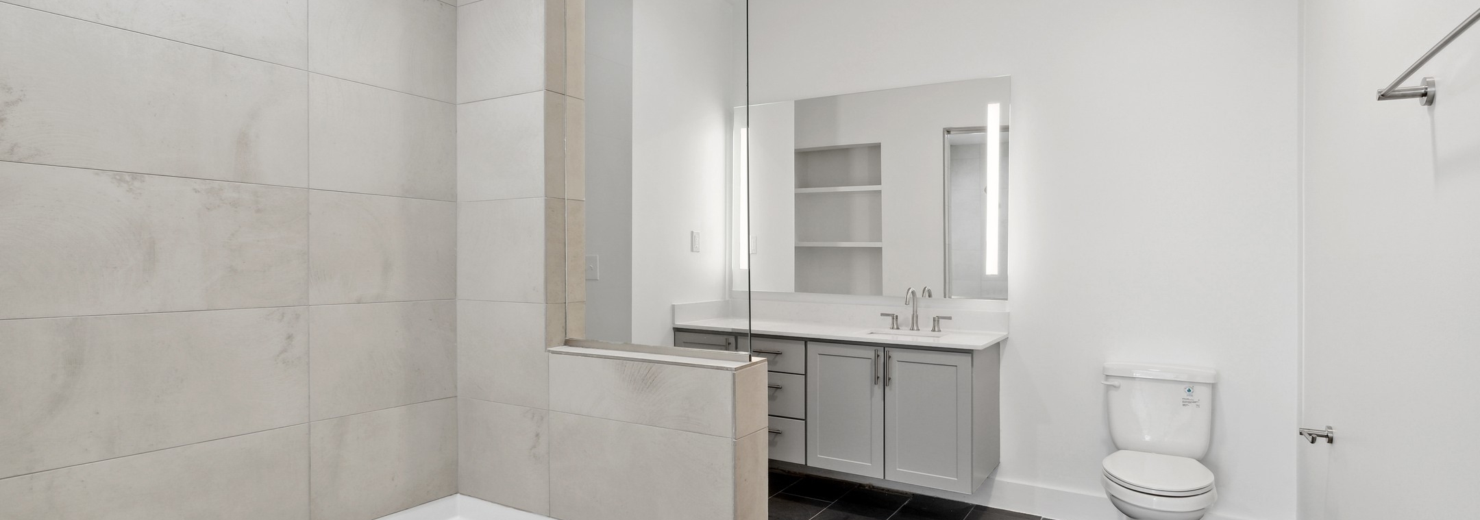 Bathroom with a cream tile tub surround and gray vanity below large mirror with black tiled flooring at AMLI Old 4th Ward