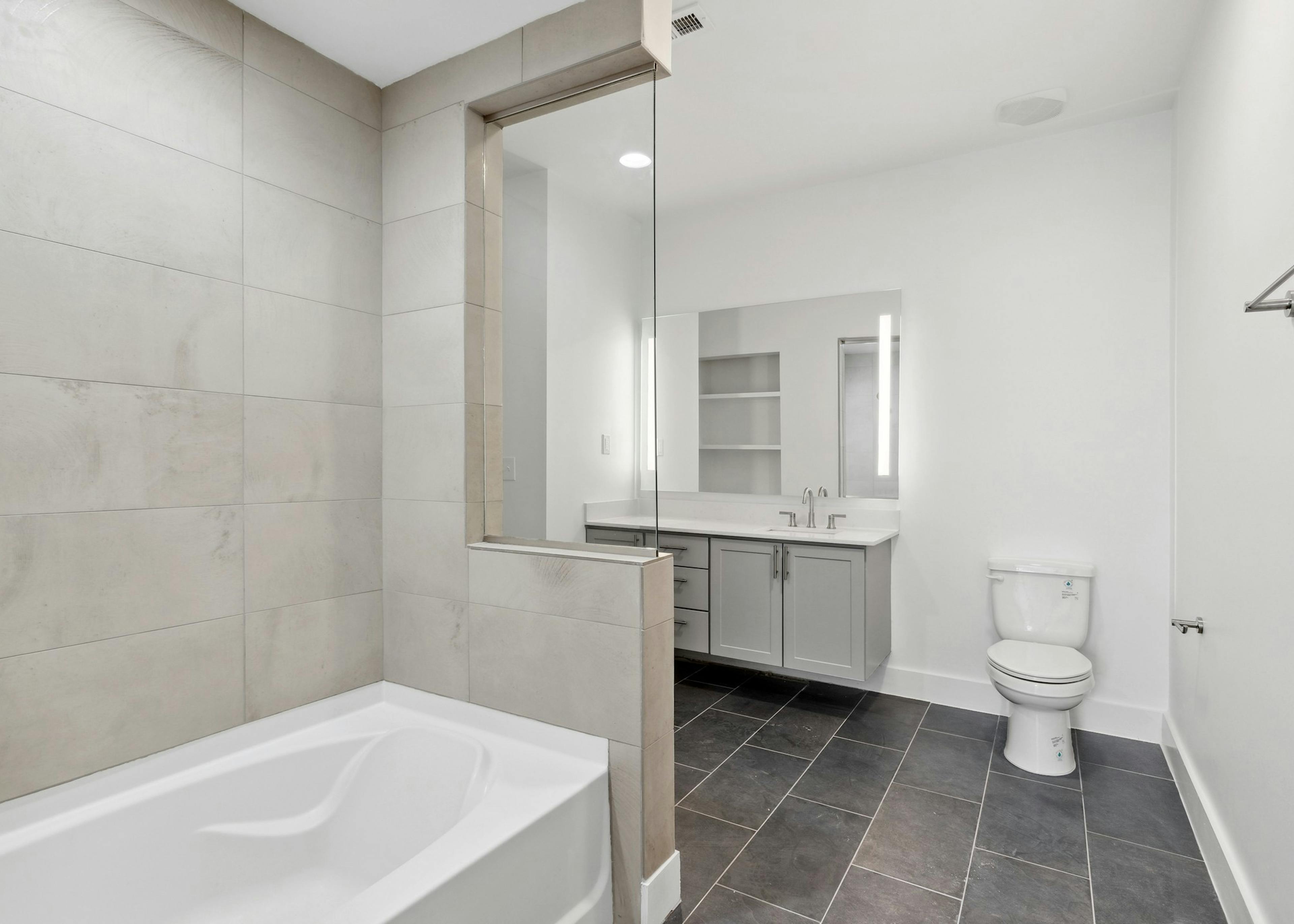 Bathroom with a cream tile tub surround and gray vanity below large mirror with black tiled flooring at AMLI Old 4th Ward