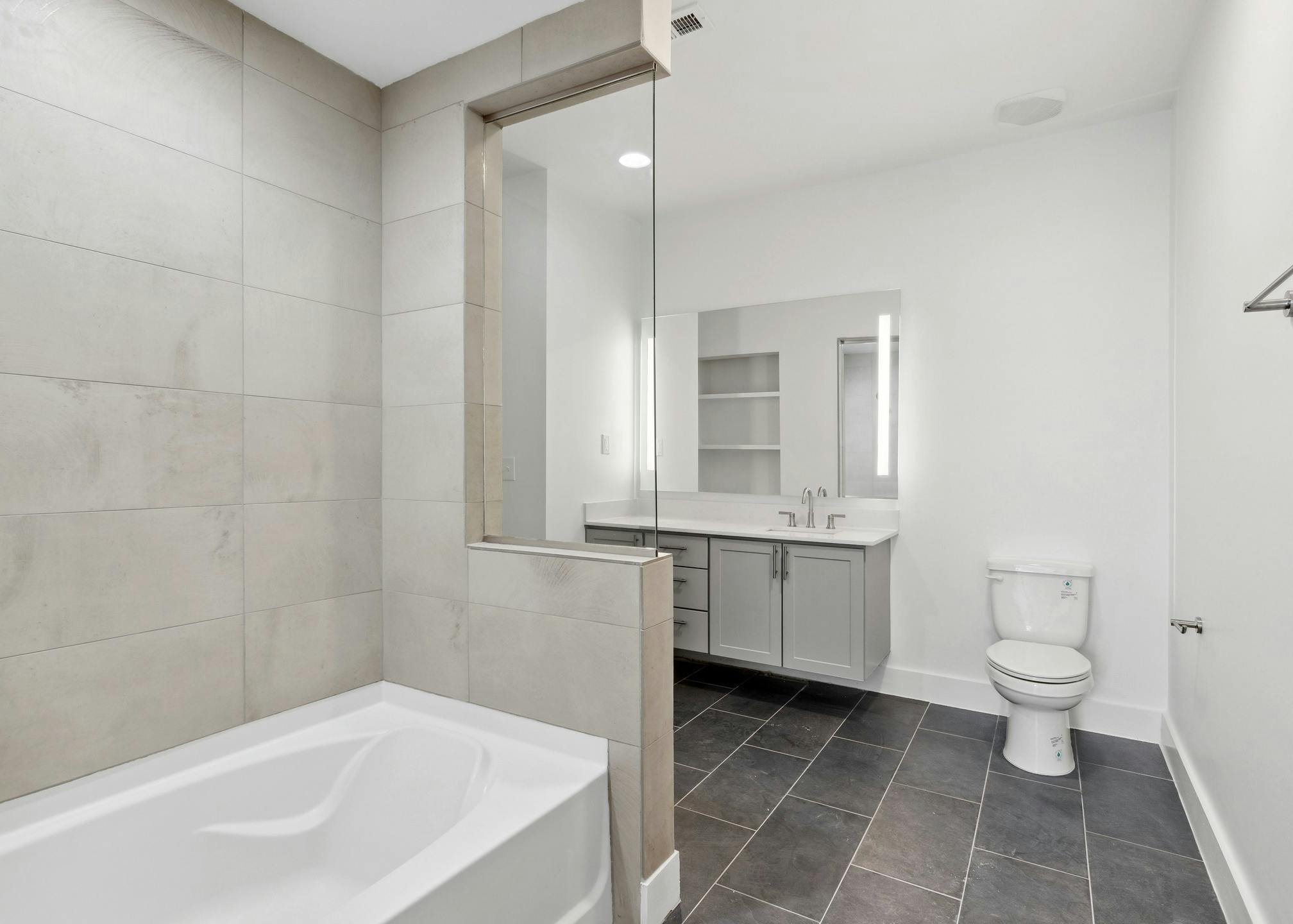 Bathroom with a cream tile tub surround and gray vanity below large mirror with black tiled flooring at AMLI Old 4th Ward