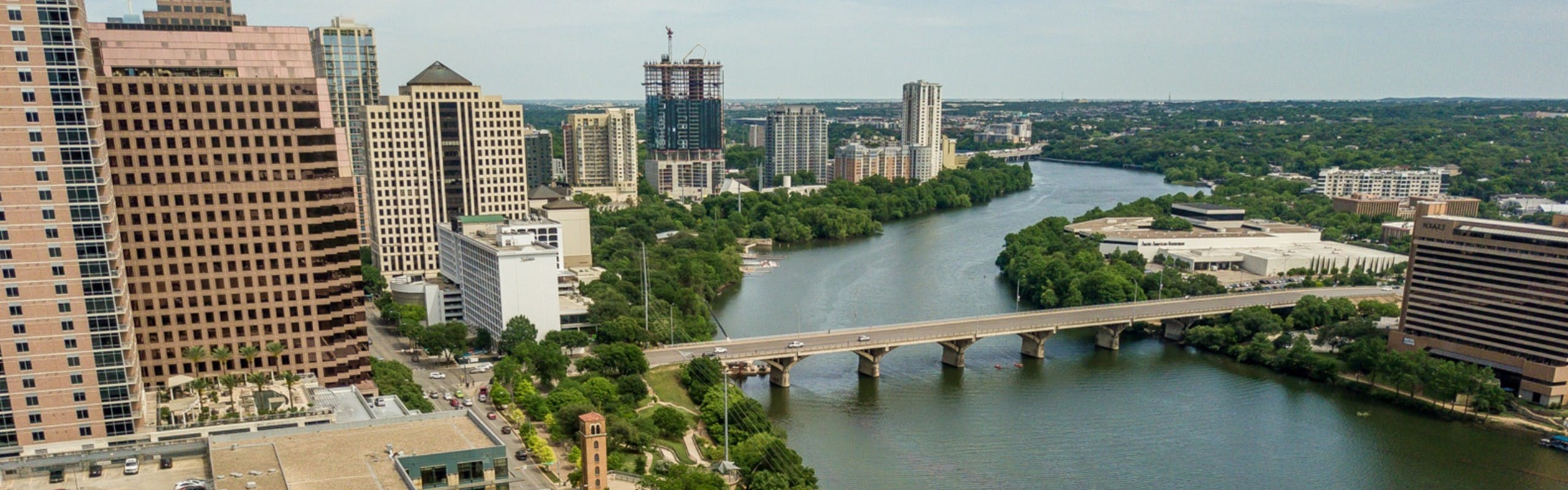 Austin city buildings with vibrant green trees separated by a long body of water with a thin bridge connecting the land