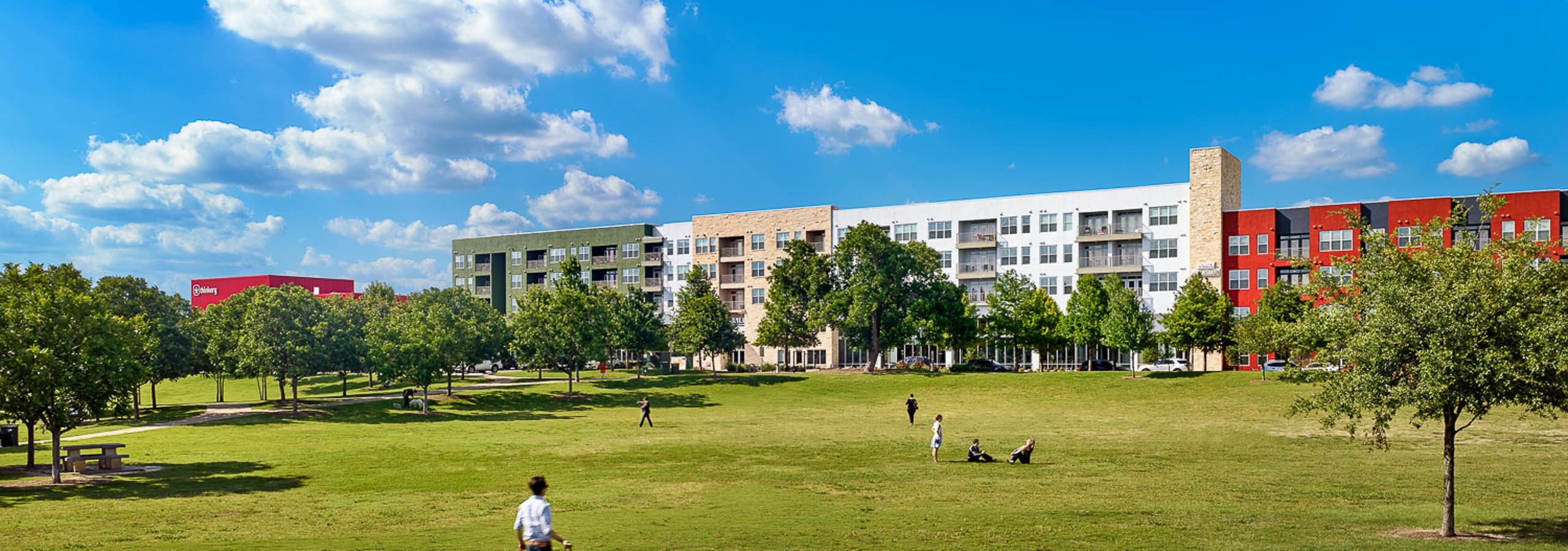 Spacious grassland area with pedestrians walking and plentiful trees with AMLI at Mueller building facade in background