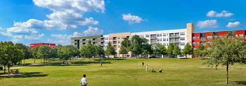 Spacious grassland area with pedestrians walking and plentiful trees with AMLI at Mueller building facade in background