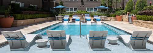 Daytime view of AMLI River Oaks pool with white lounge chairs in the pool with blue pillows attached and surrounding greenery