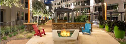 Fire pit on pavement at AMLI on Aldrich with one blue and one red chair surrounded by potted plants and greenery at night