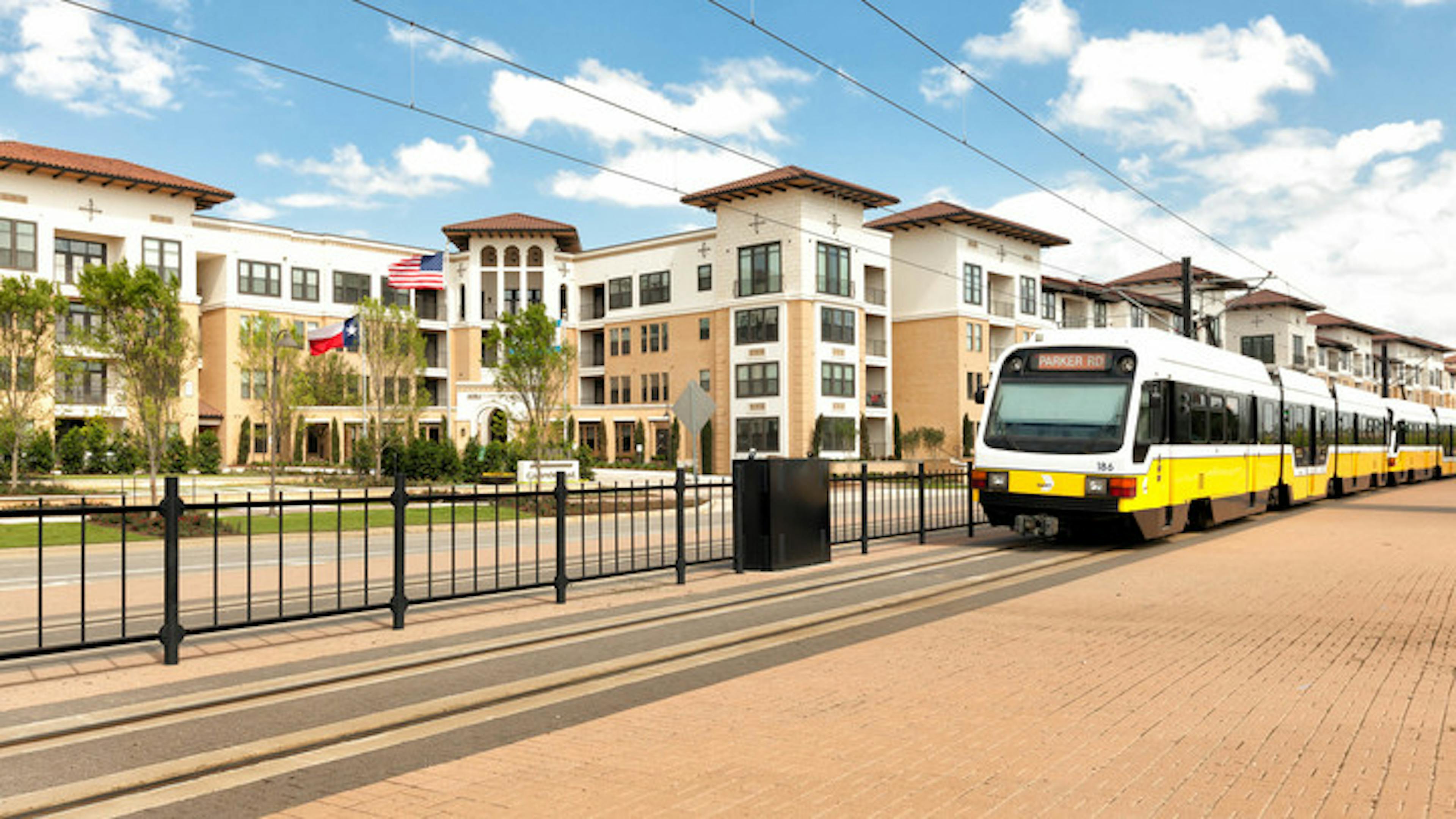 Daytime view of light rail train on tracks along fence traveling in front of the AMLI Campion Trail apartment building