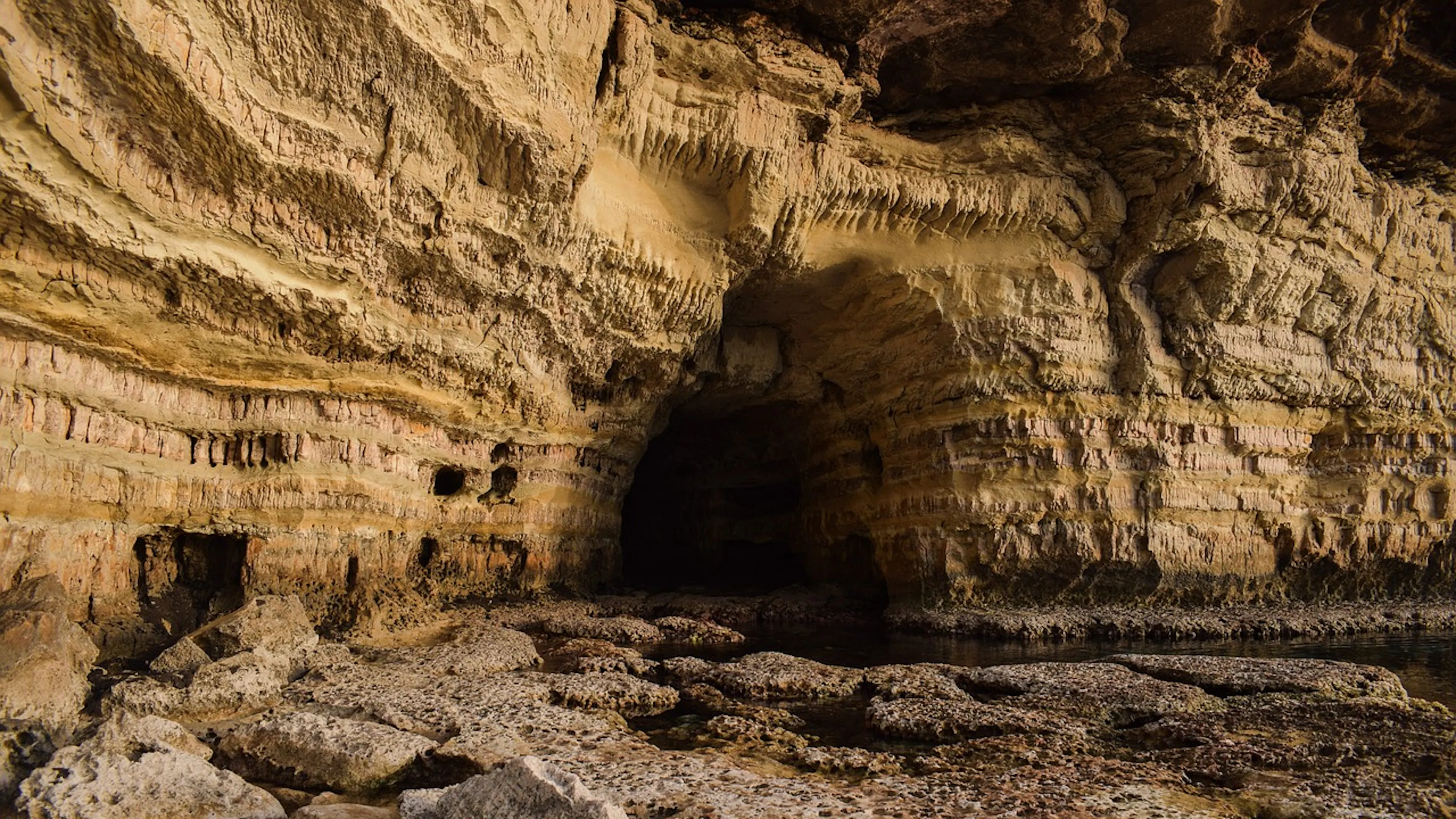 A cave entrance in a limestone rock cliff face surrounded by some water