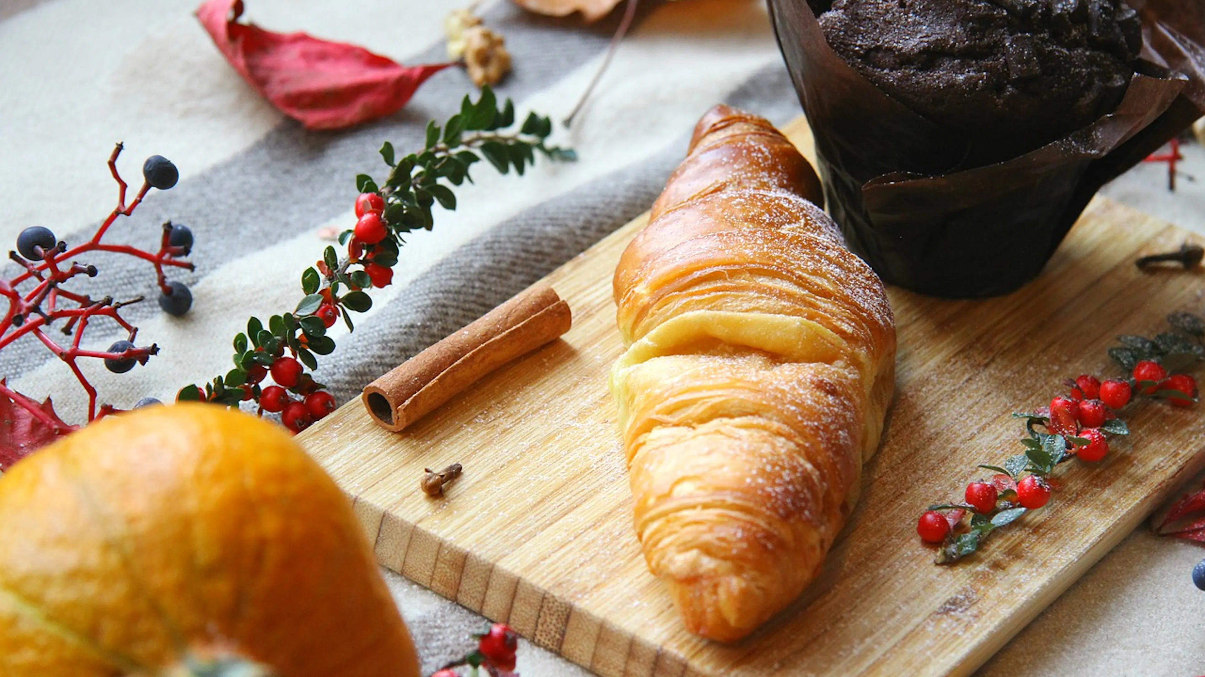 A croissant and a chocolate muffin on a small wooden cutting board surrounded by a pumpkin and some sprigs of holly