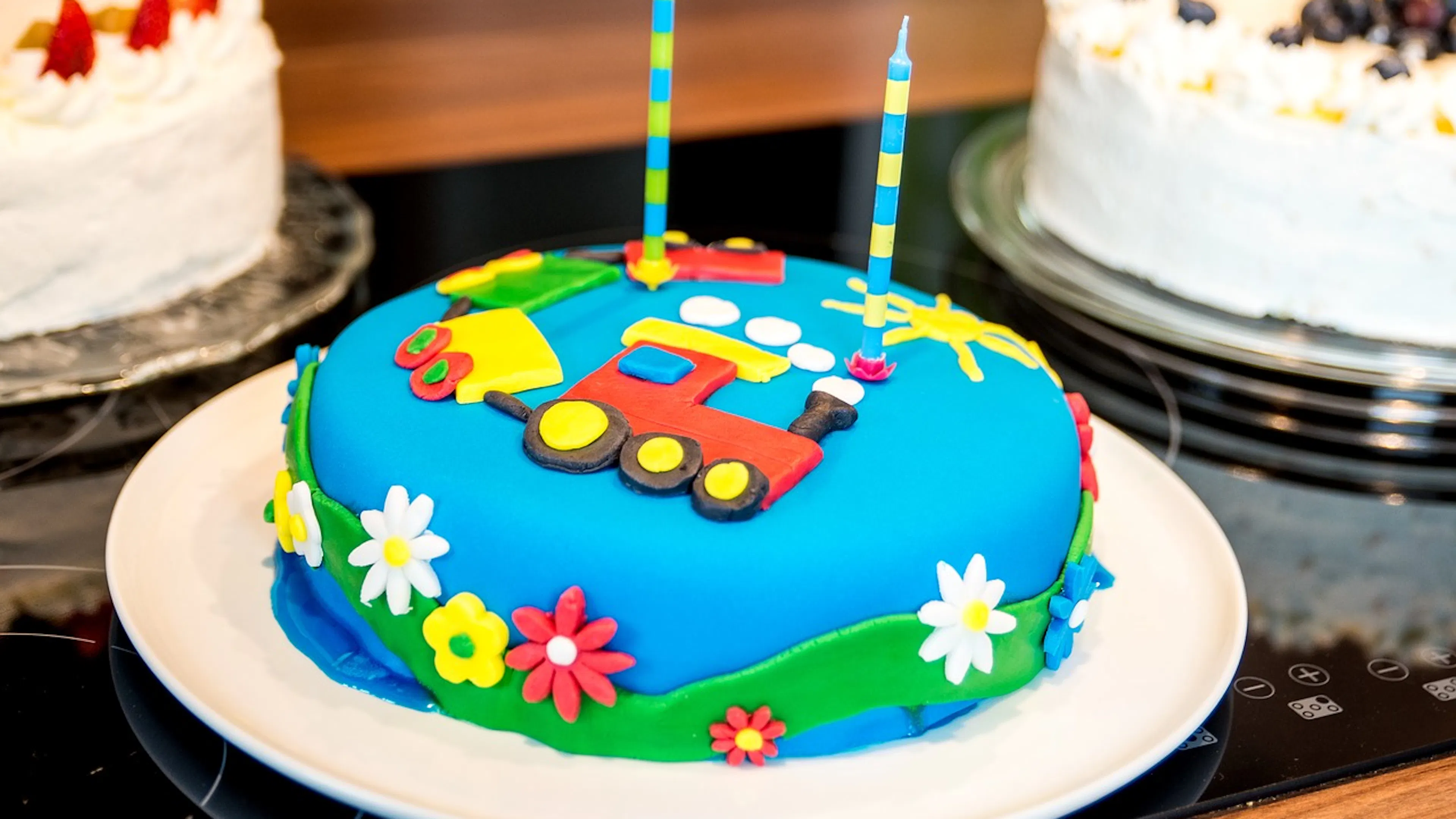 Blue birthday cake with tractor and flower with two blue and yellow candles on top on a white plate beside other cakes