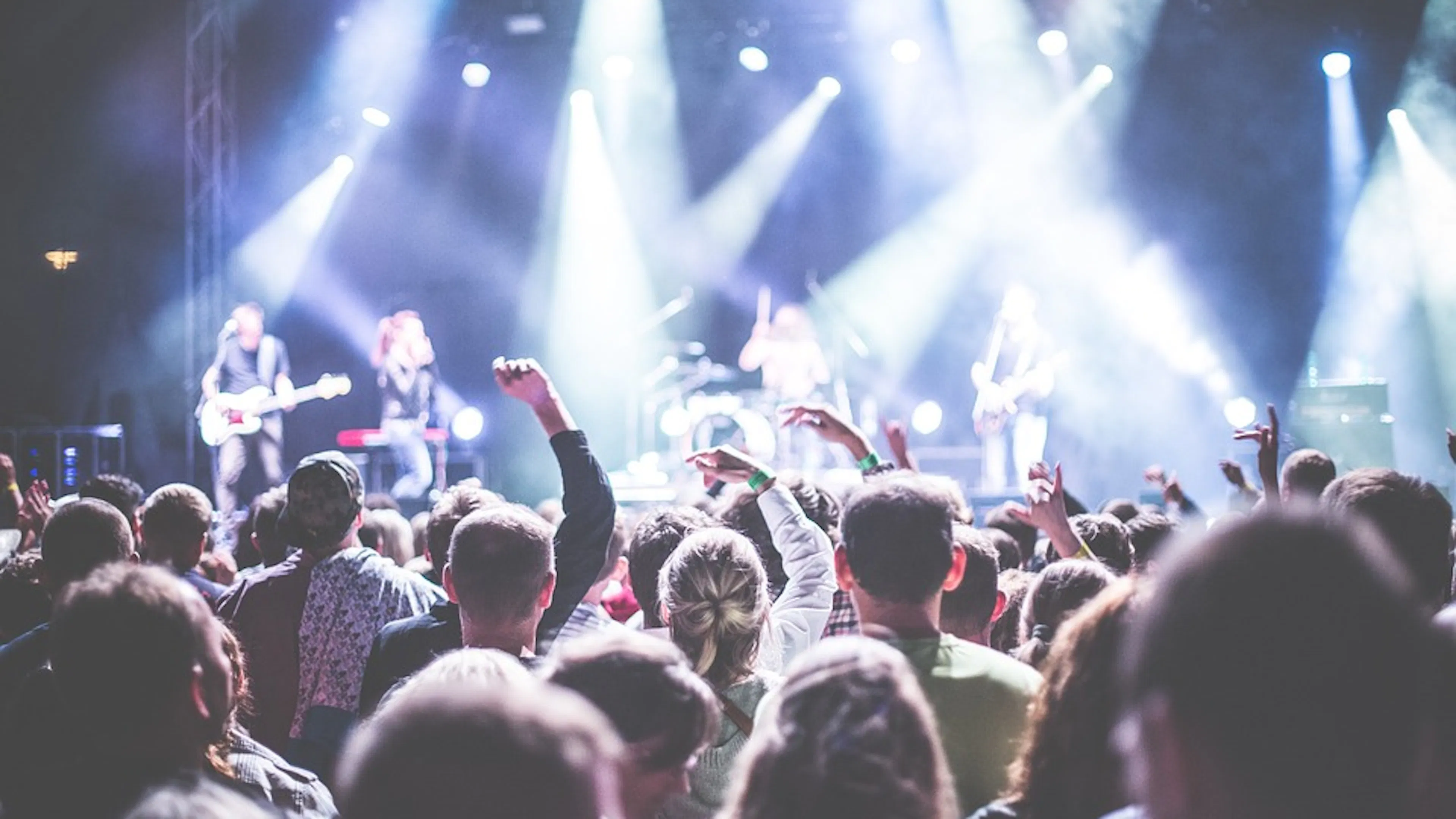 A crowd of people facing a concert stage with bright spotlights lighting up the audiences excited hands in the air