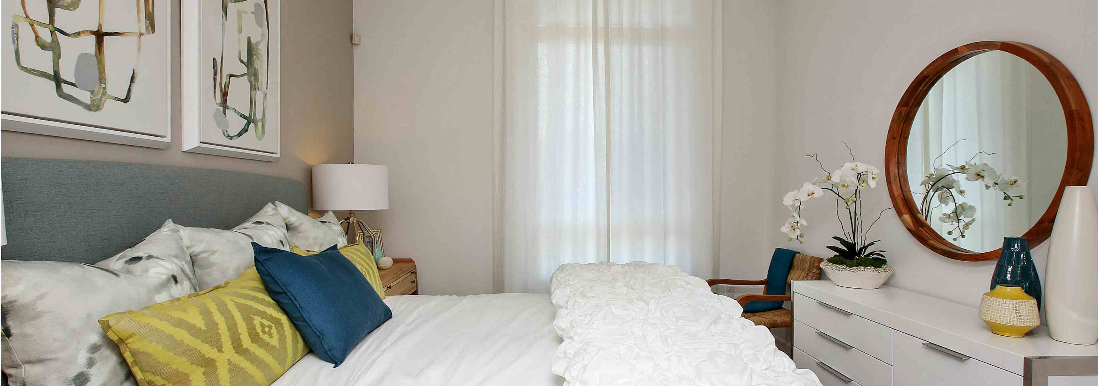 Interior view of AMLI Uptown Orange furnished apartment bedroom with king bed, dresser and white curtains over large window