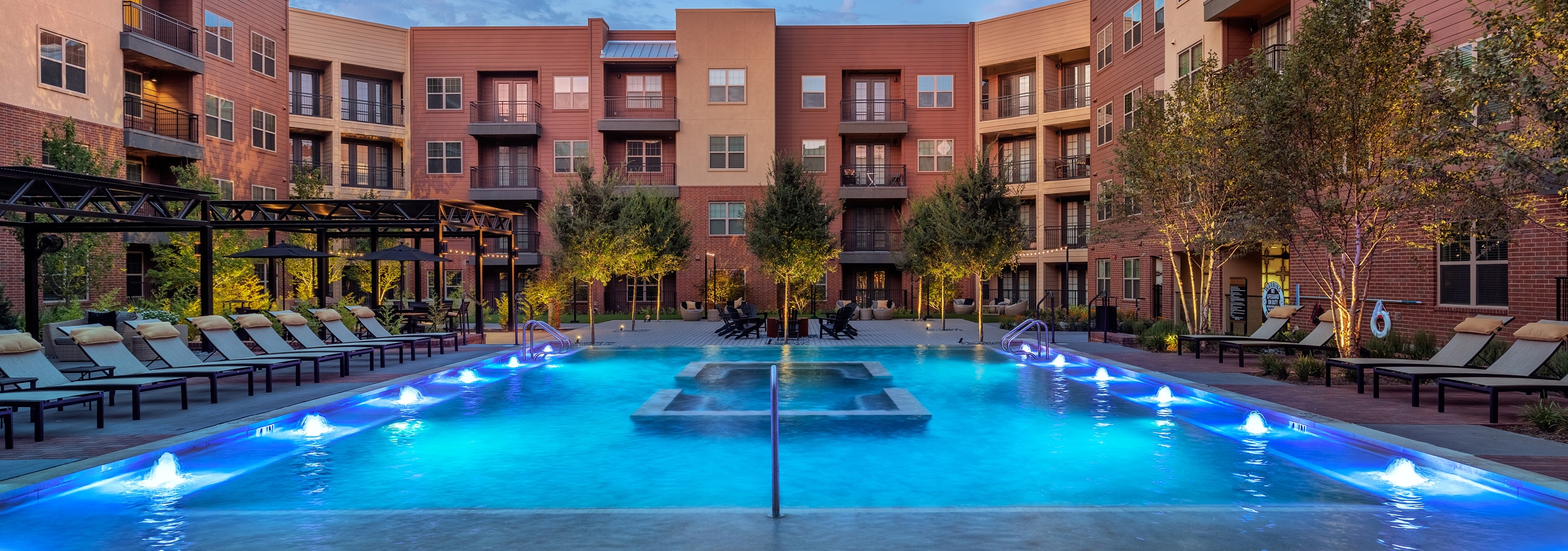 AMLI Grapevine swimming pool at dusk with pool lights and surrounded by green trees and light gray lounge seating