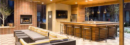 Outdoor brick fireplace at AMLI West Plano with a yellow and green couch and barstools at a long counter in the background