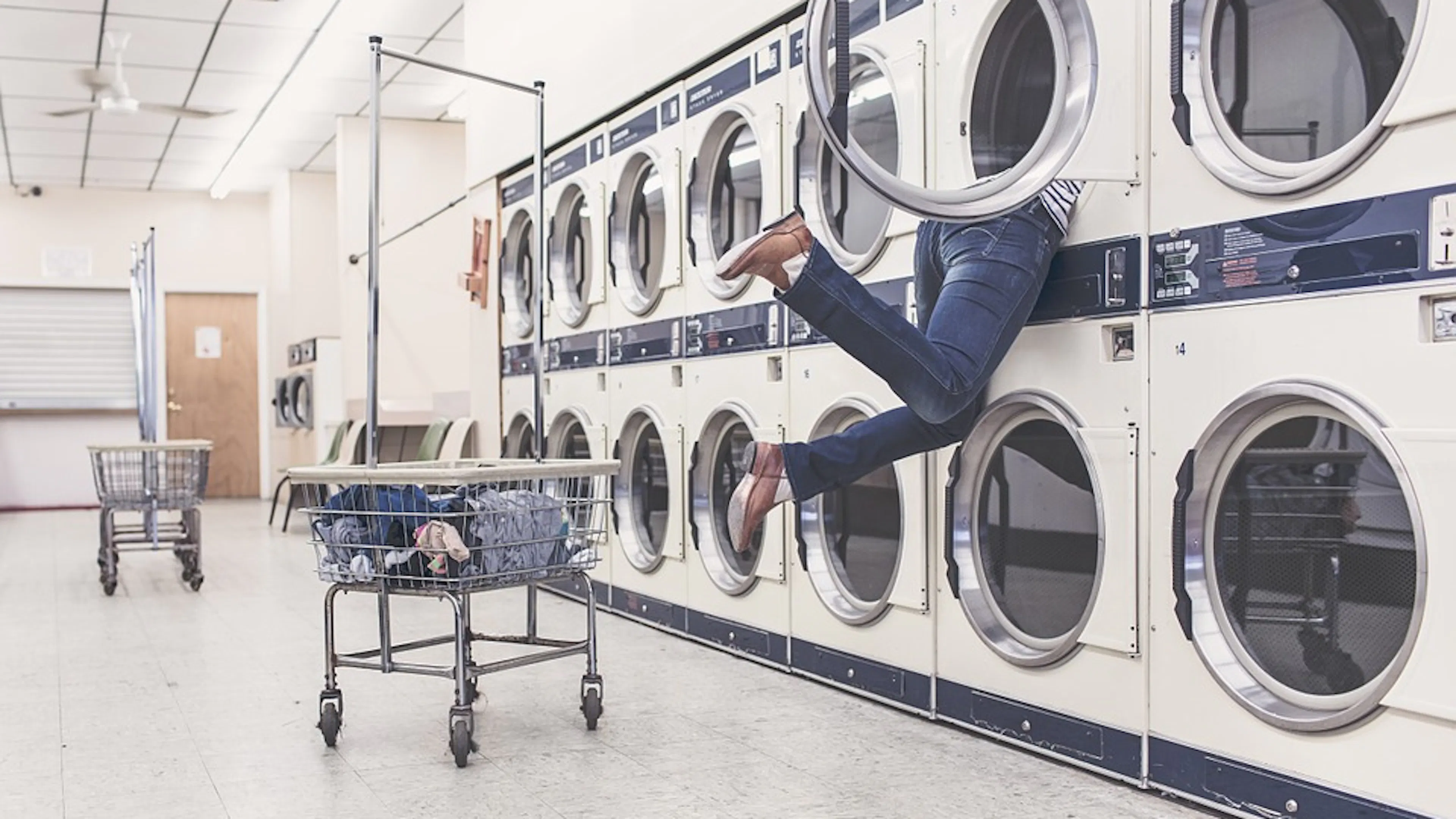 a woman reaching into a tumble dryer in an empty laundromat with her feet off the ground