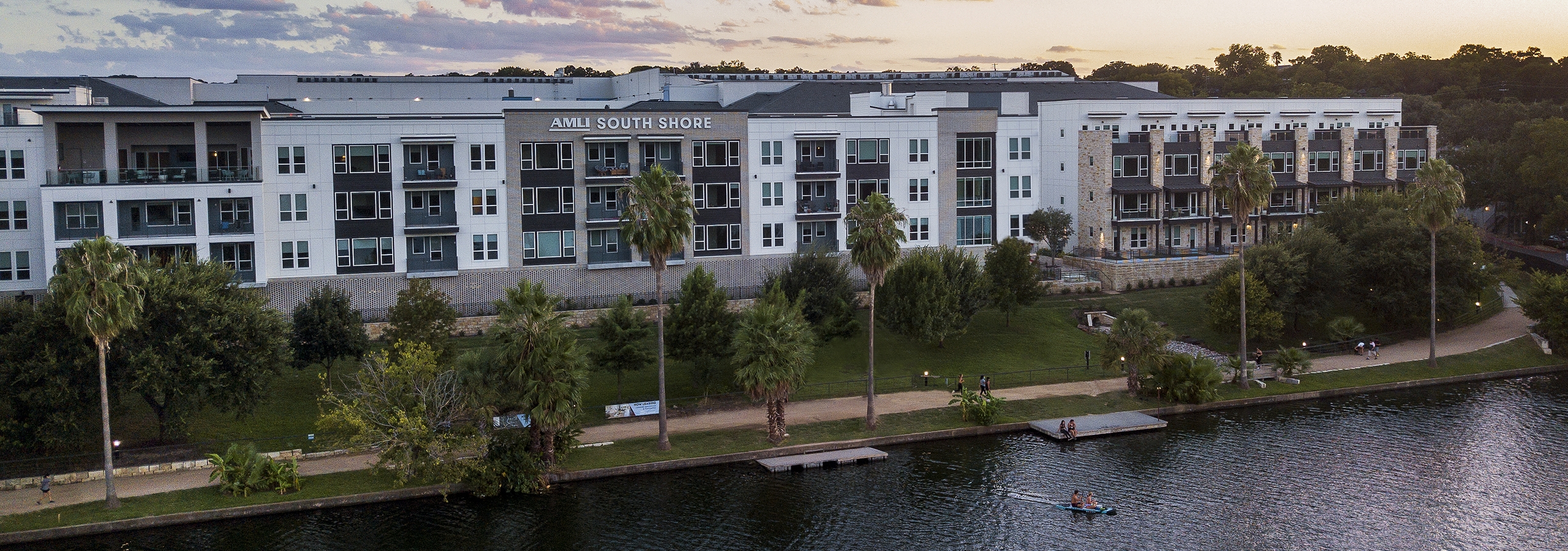 An aerial view at sunset of AMLI South Shore exterior façade with people kayaking in the lake and enjoying the walking trail