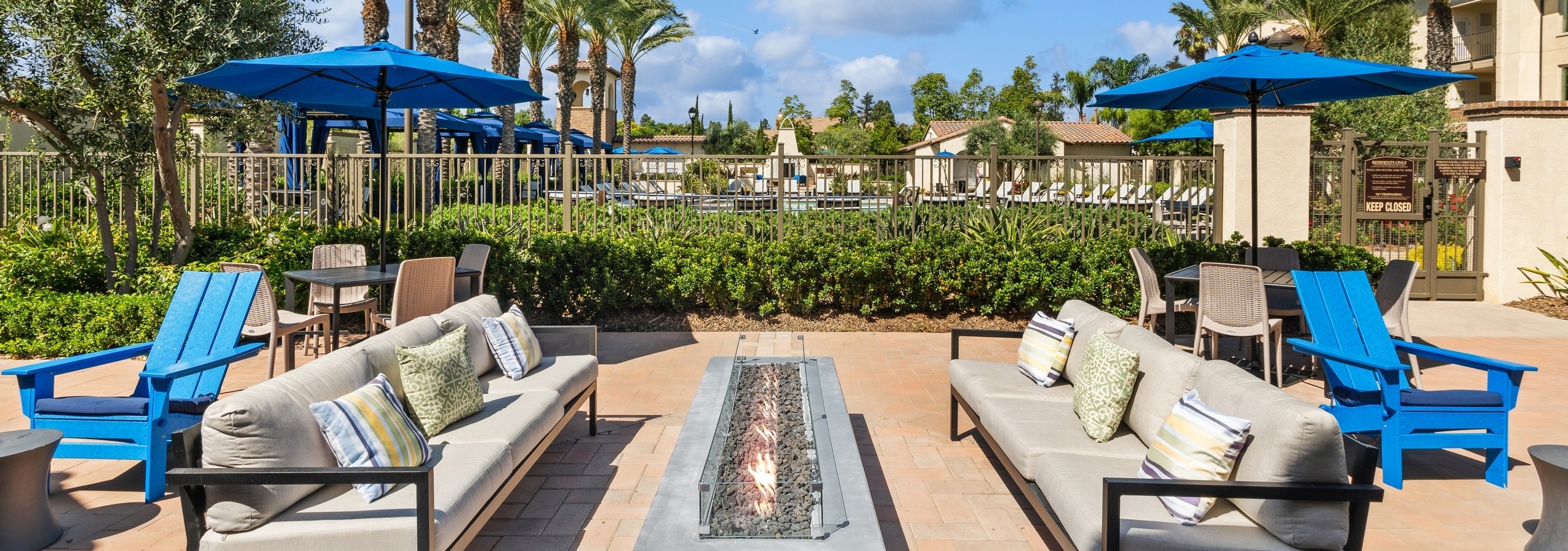 Exterior view of couch and Adirondack seating at AMLI Spanish Hills surrounding a long lit firepit next to the pool fence