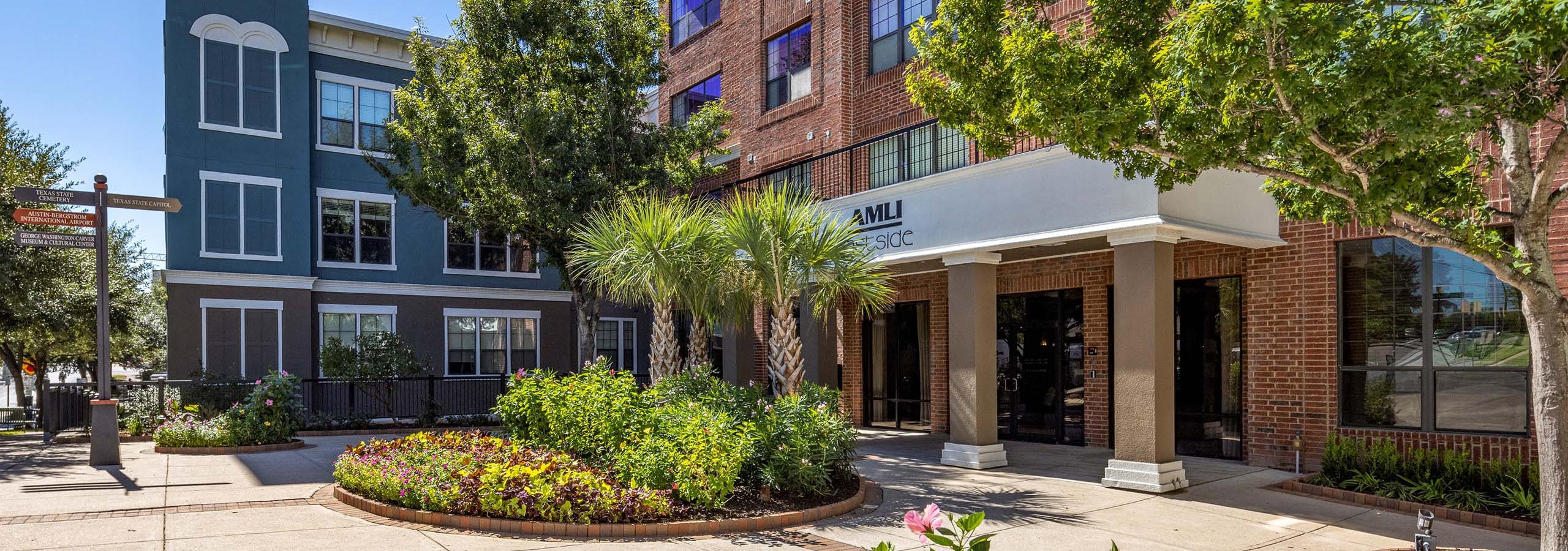 Sunny view of AMLI Eastside apartments main entrance surrounded by green palm trees and blooming colorful flowers