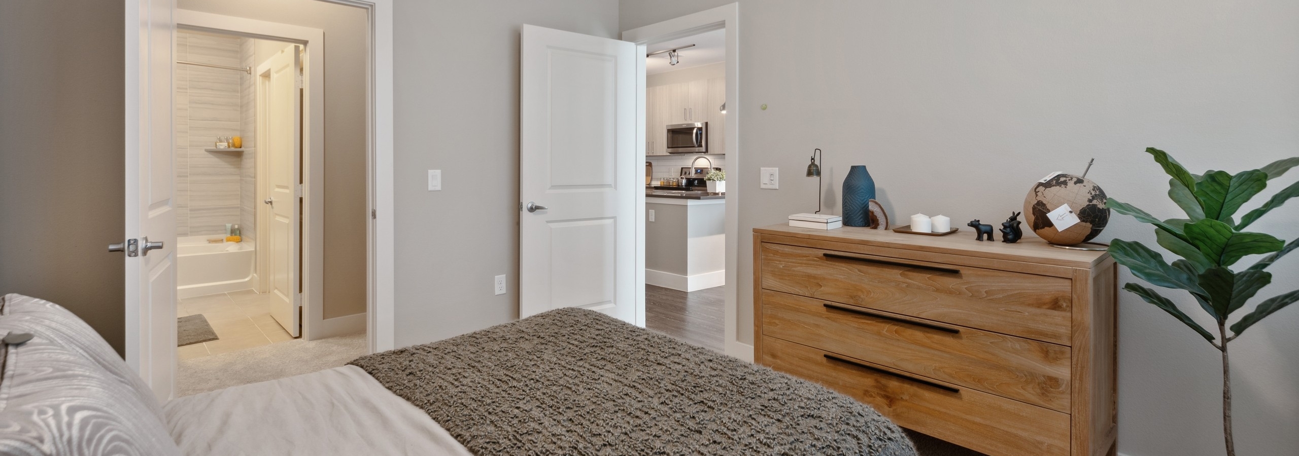 Bedroom at AMLI Dry Creek apartments with gray and brown linens and wood dresser and plant with view to bathroom
