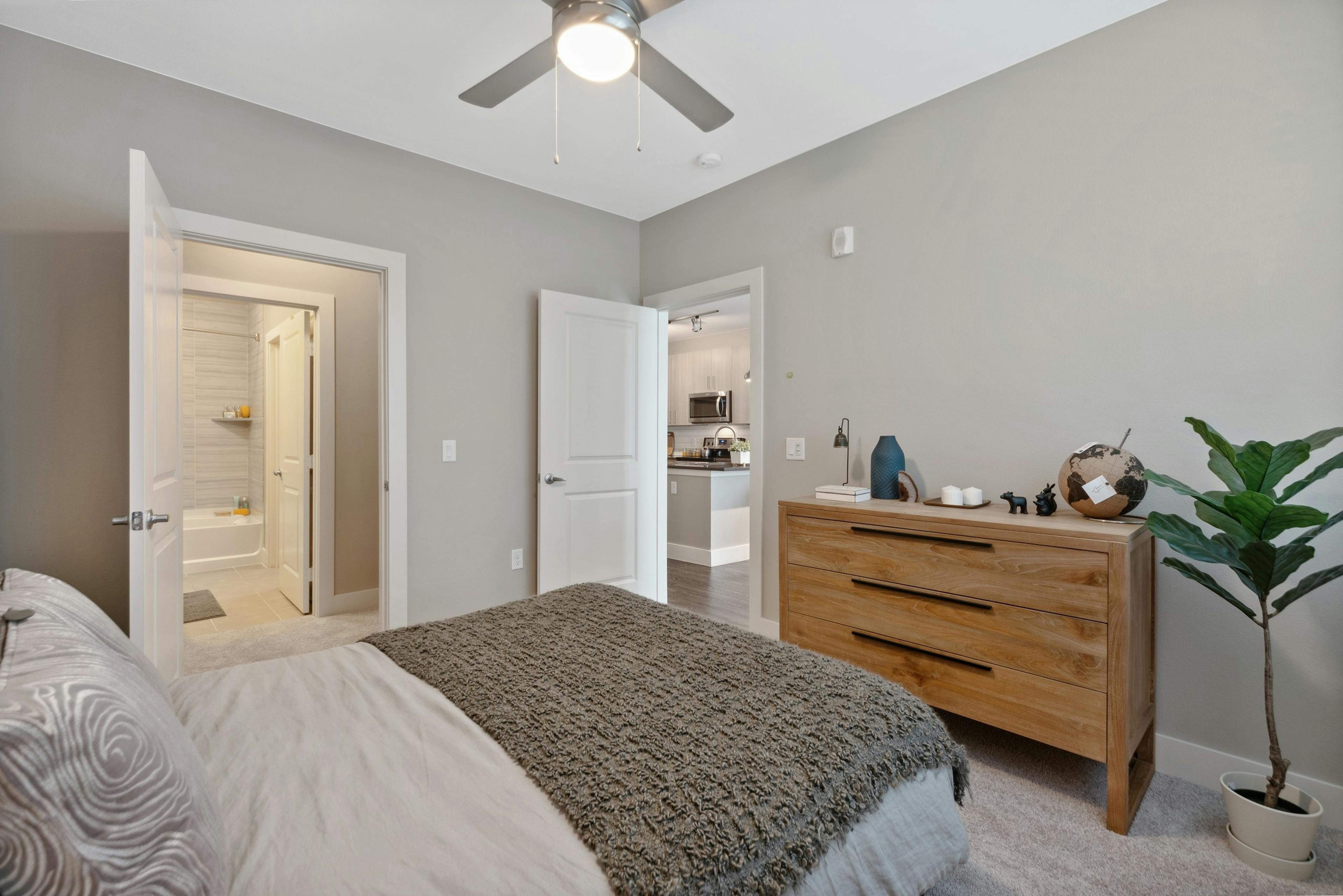 Bedroom at AMLI Dry Creek apartments with gray and brown linens and wood dresser and plant with view to bathroom