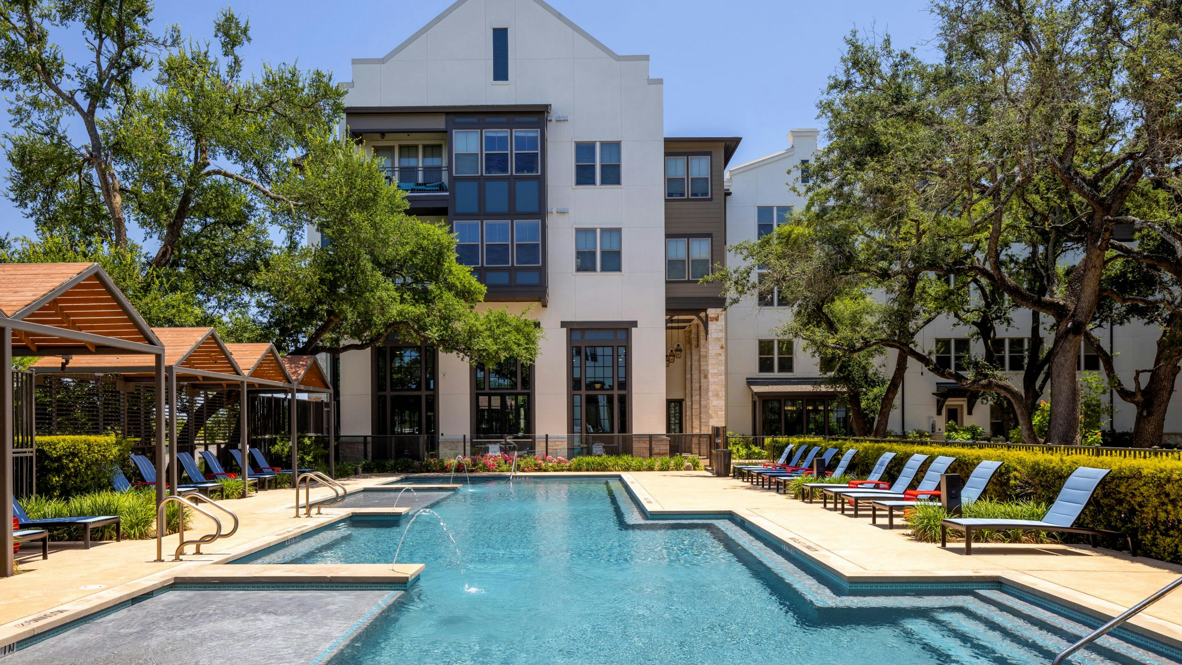 Daytime view of swimming pool at AMLI Lakeline with wood cabanas and blue lounge chairs and red tables and lush greenery