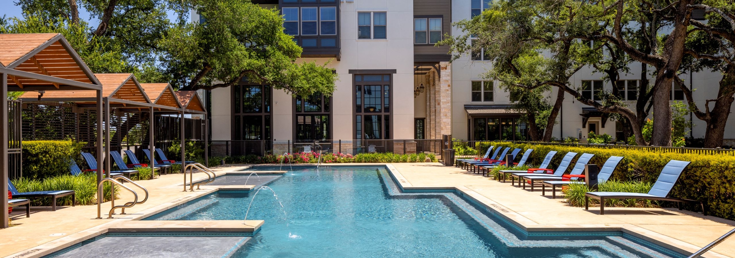 Daytime view of swimming pool at AMLI Lakeline with wood cabanas and blue lounge chairs and red tables and lush greenery