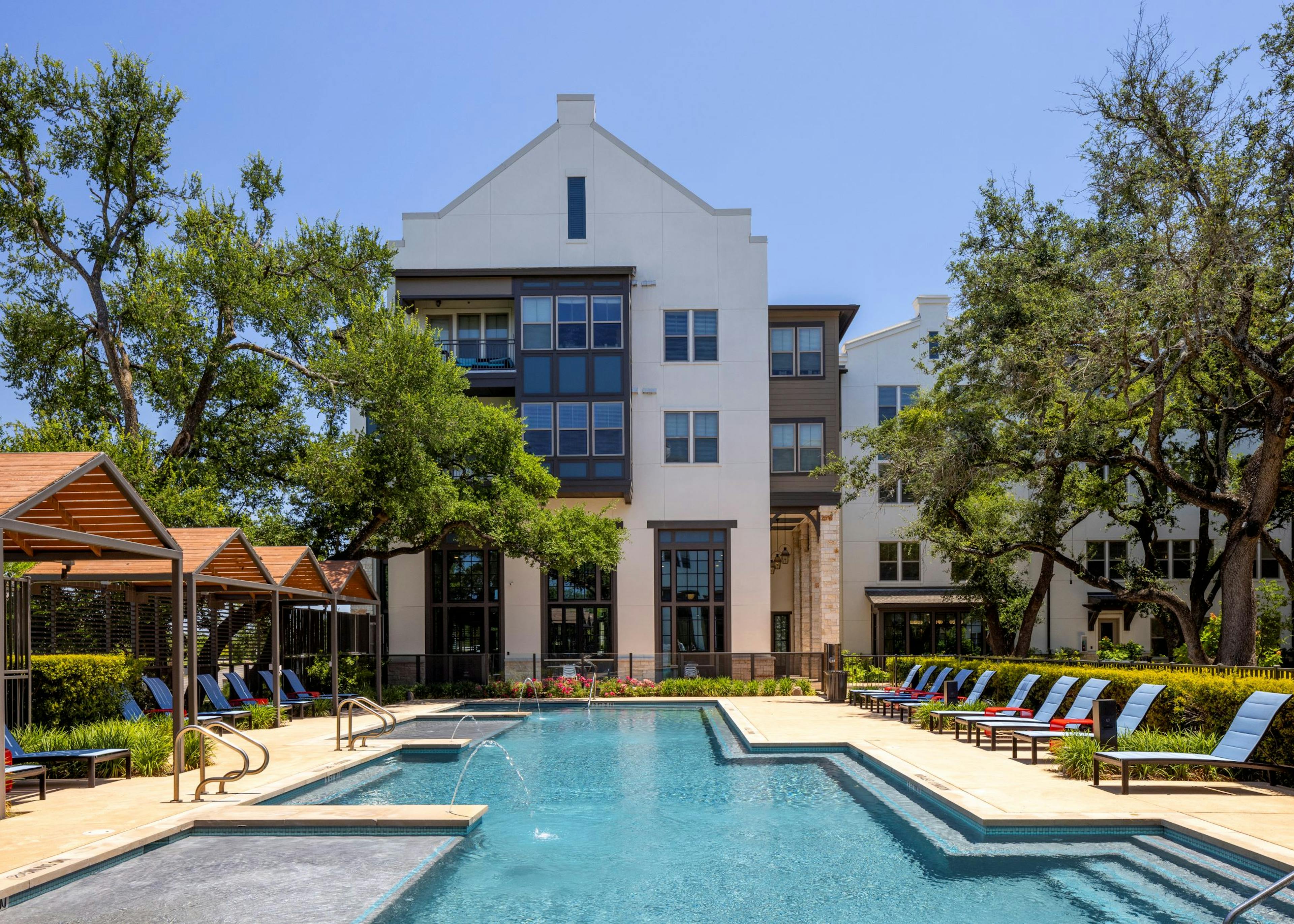 Daytime view of swimming pool at AMLI Lakeline with wood cabanas and blue lounge chairs and red tables and lush greenery