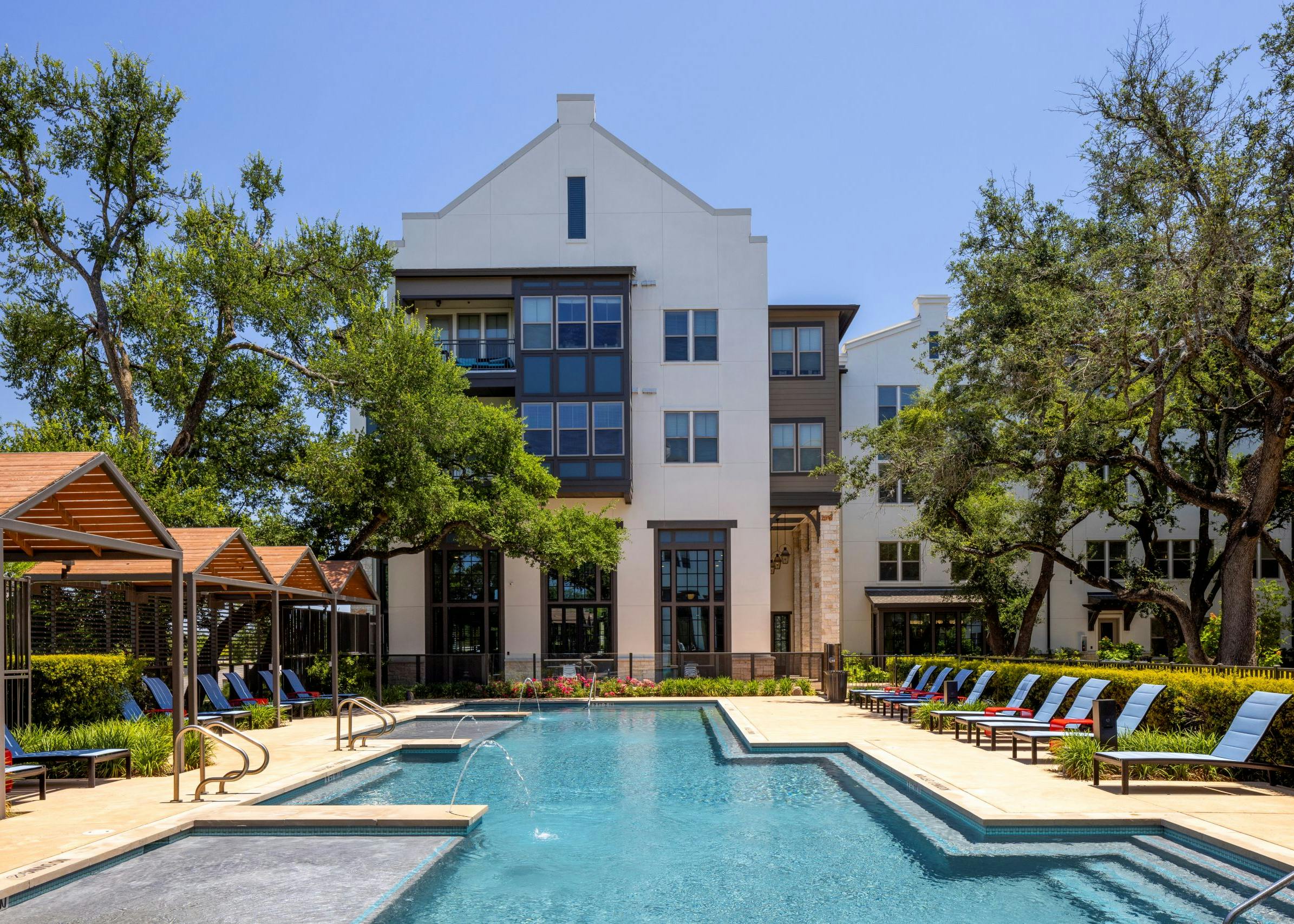Daytime view of swimming pool at AMLI Lakeline with wood cabanas and blue lounge chairs and red tables and lush greenery