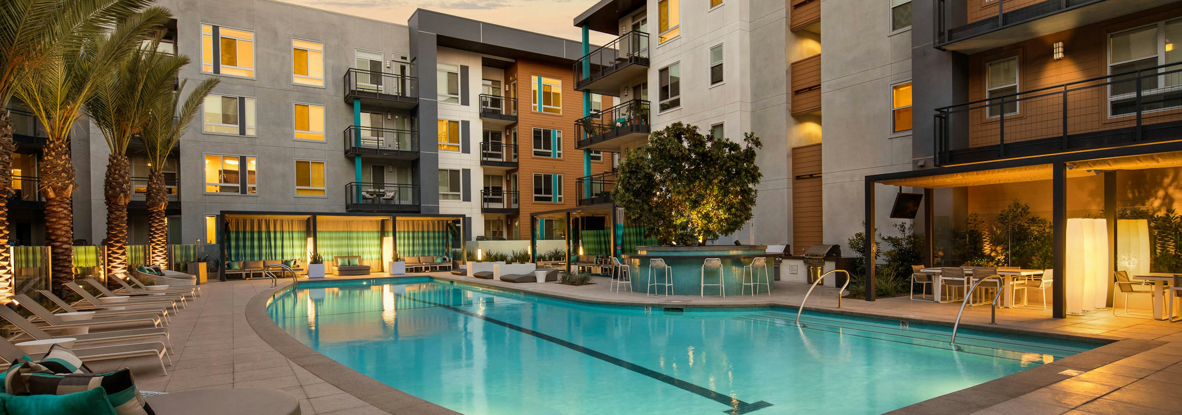 Dusk view of AMLI Uptown Orange swimming pool area with lounge seating, cabanas with TVs and seating and lights on palm trees