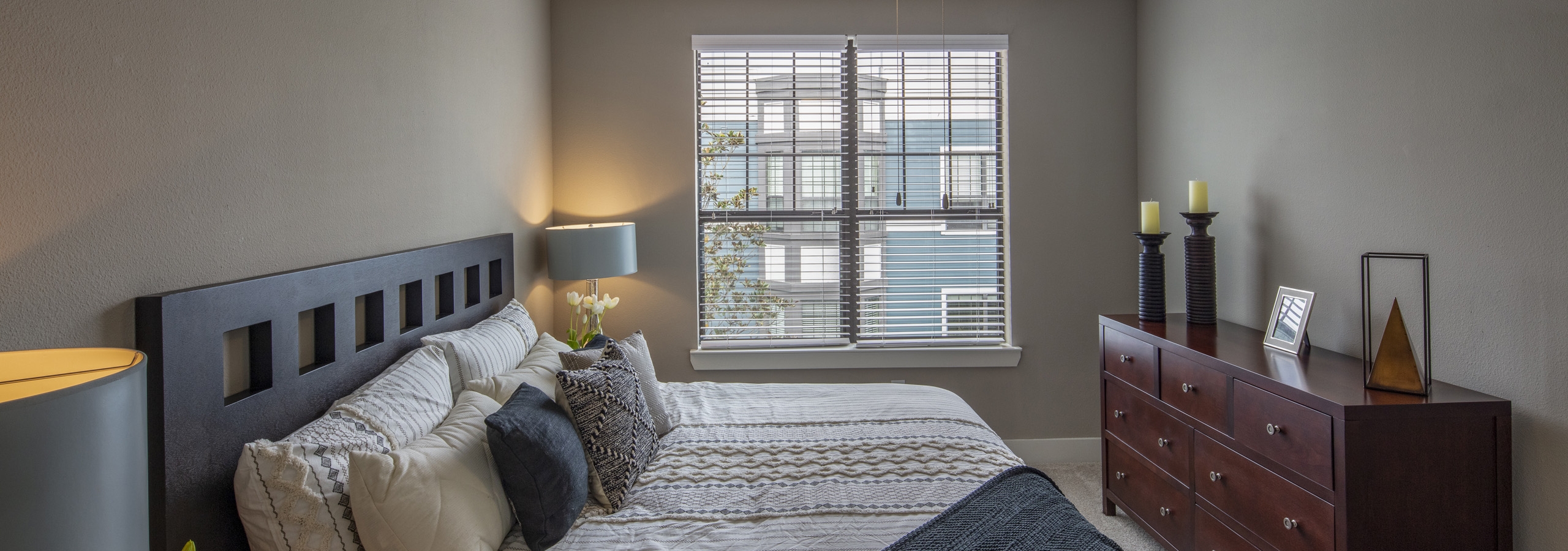 Day view of carpeted bedroom at AMLI Eastside apartments with large window with blinds and a Queen sized bed and wood dresser