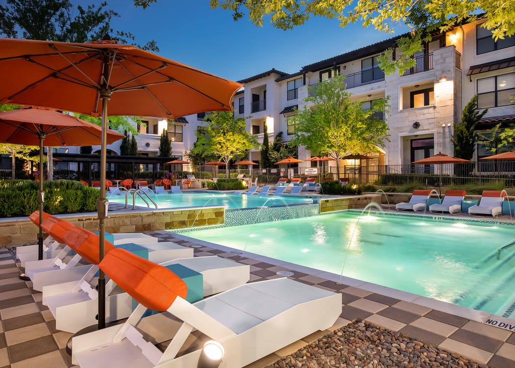 Pool at AMLI on Riverside at night with orange umbrellas, white lounge chairs and vibrant trees surrounding the water