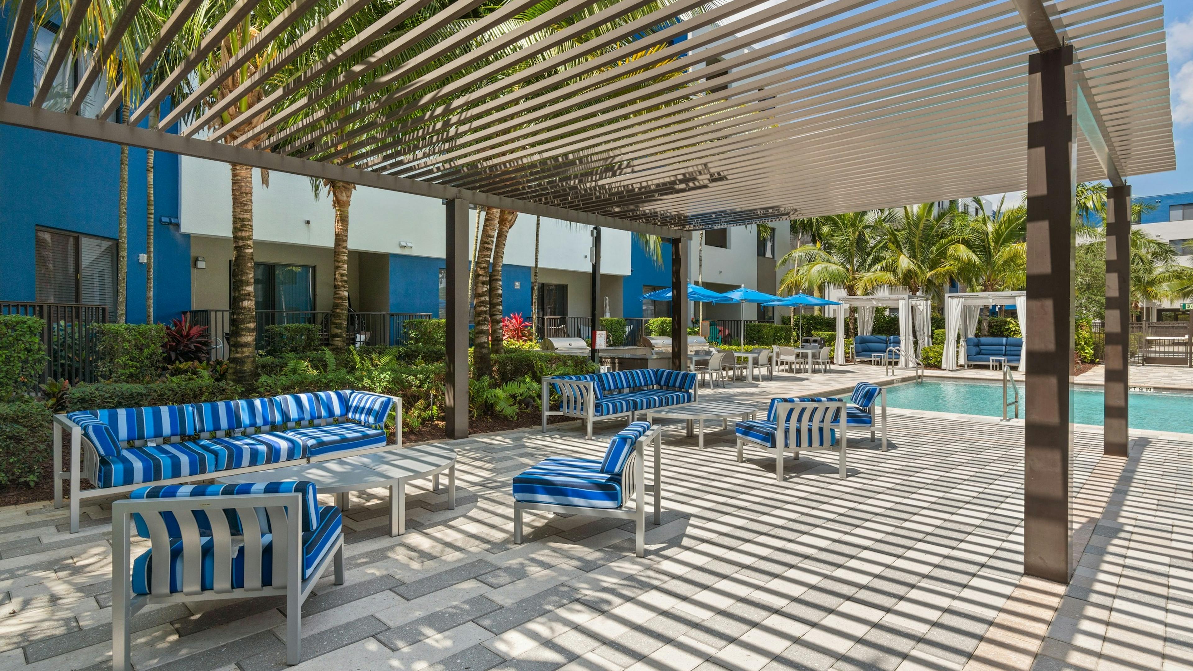 The poolside lounge at AMLI 8800 with pergola and blue striped sofas and chairs with oval tables and tropical landscaping 