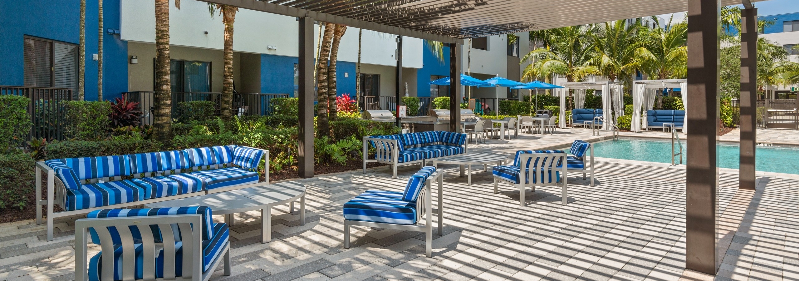 The poolside lounge at AMLI 8800 with pergola and blue striped sofas and chairs with oval tables and tropical landscaping