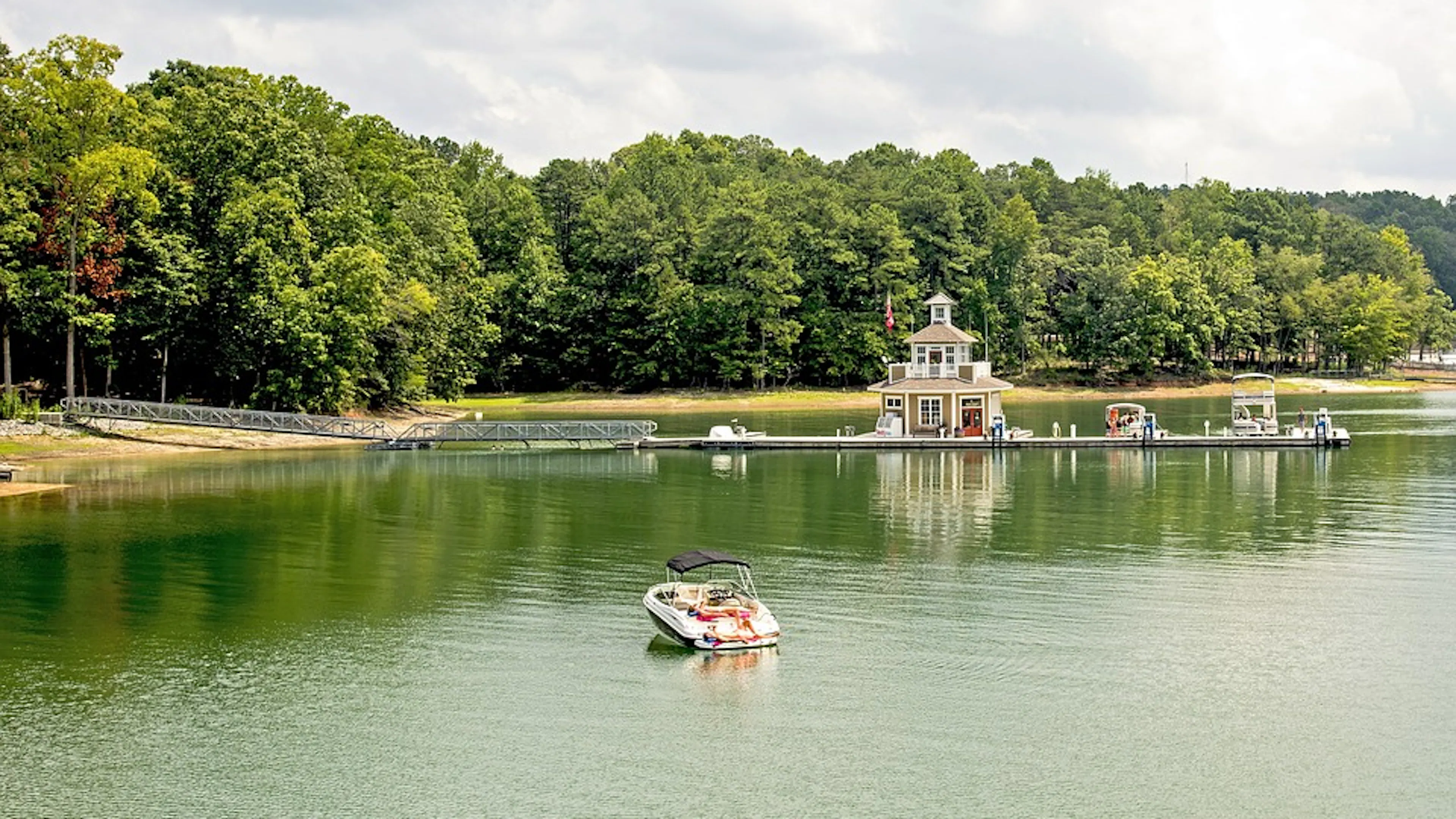 A pontoon boat heading to the grassy shore of a calm lake on a sunny day