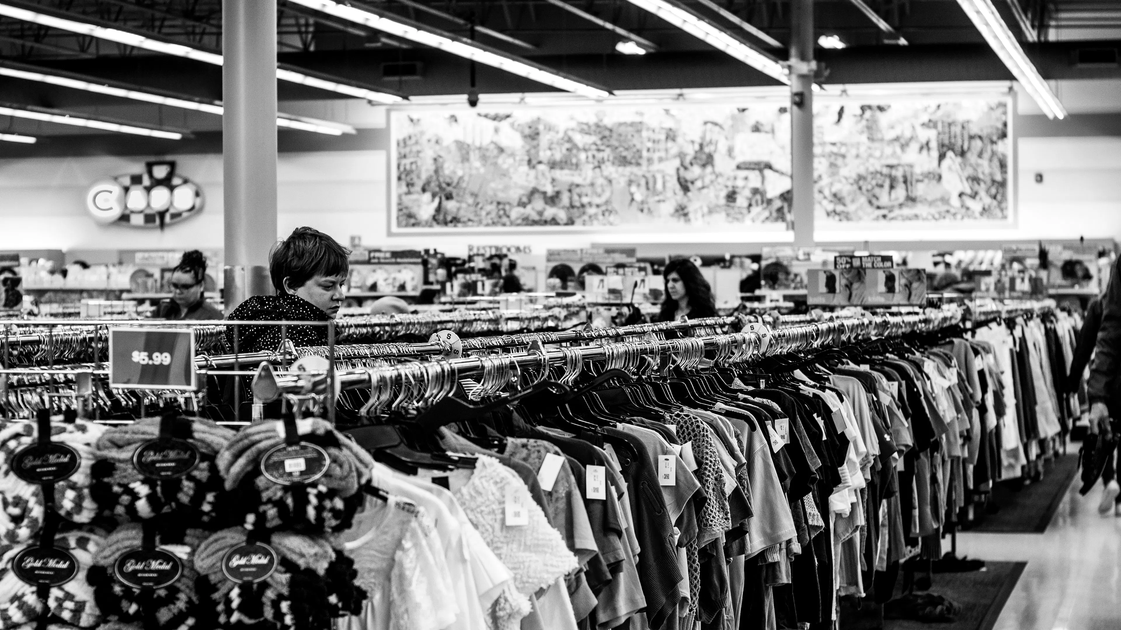 A black and white image of a woman browsing through a thrift store in between long racks of T shirts
