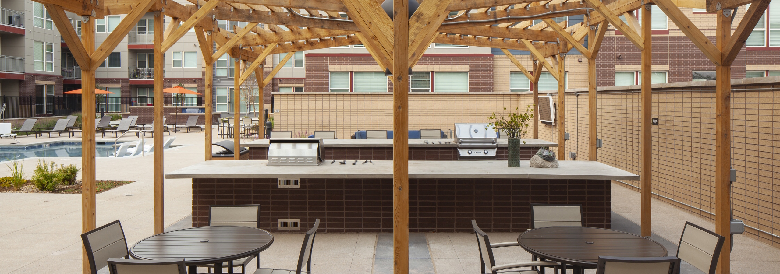 Exterior view of a grilling area at AMLI Littleton Village pool with 2 grills and 2 tables with chairs and a large wood canopy